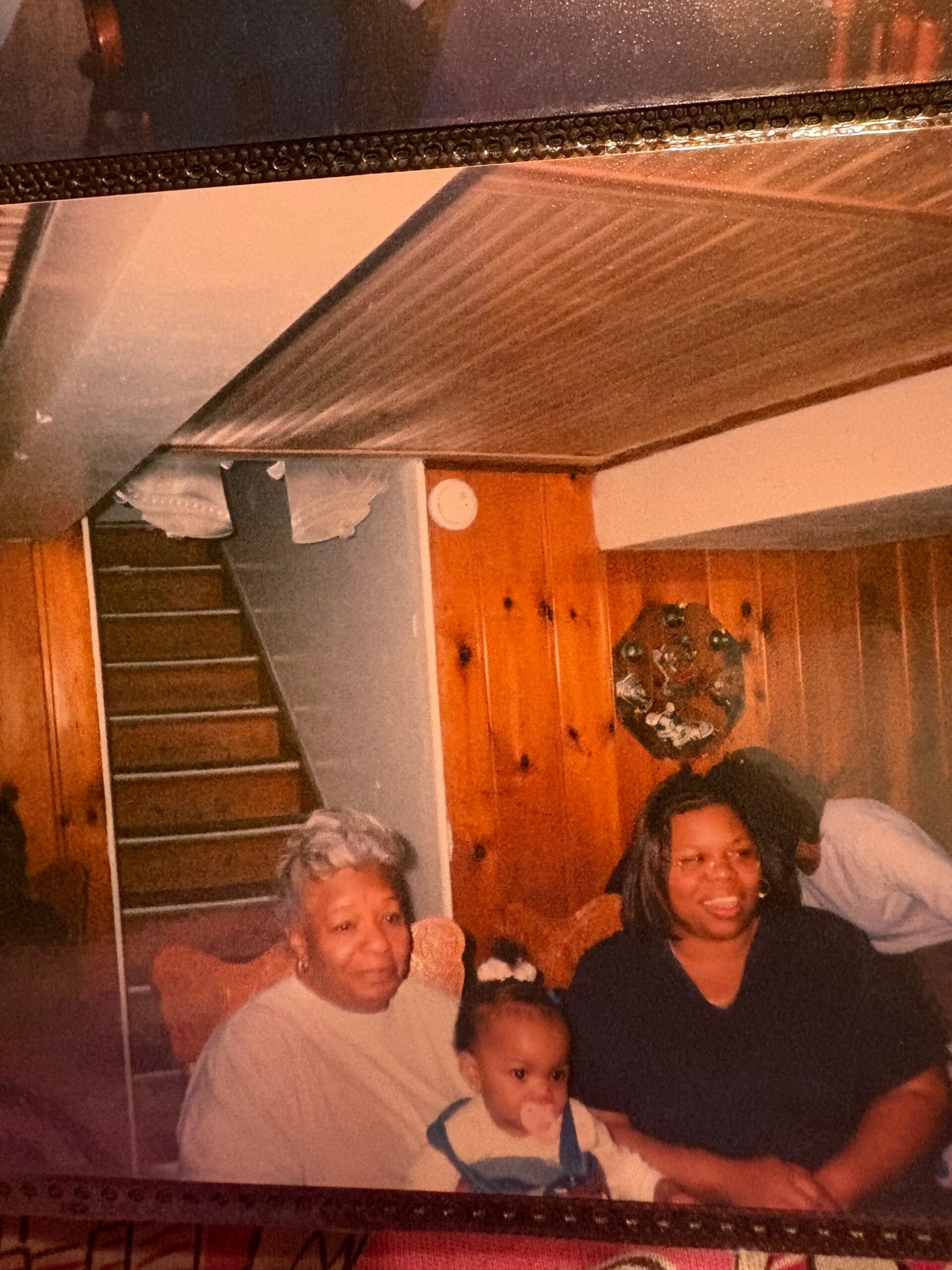 Family group photo in a wood-paneled room; two adults and a baby with a pacifier sit together, smiling, with a staircase in the background.