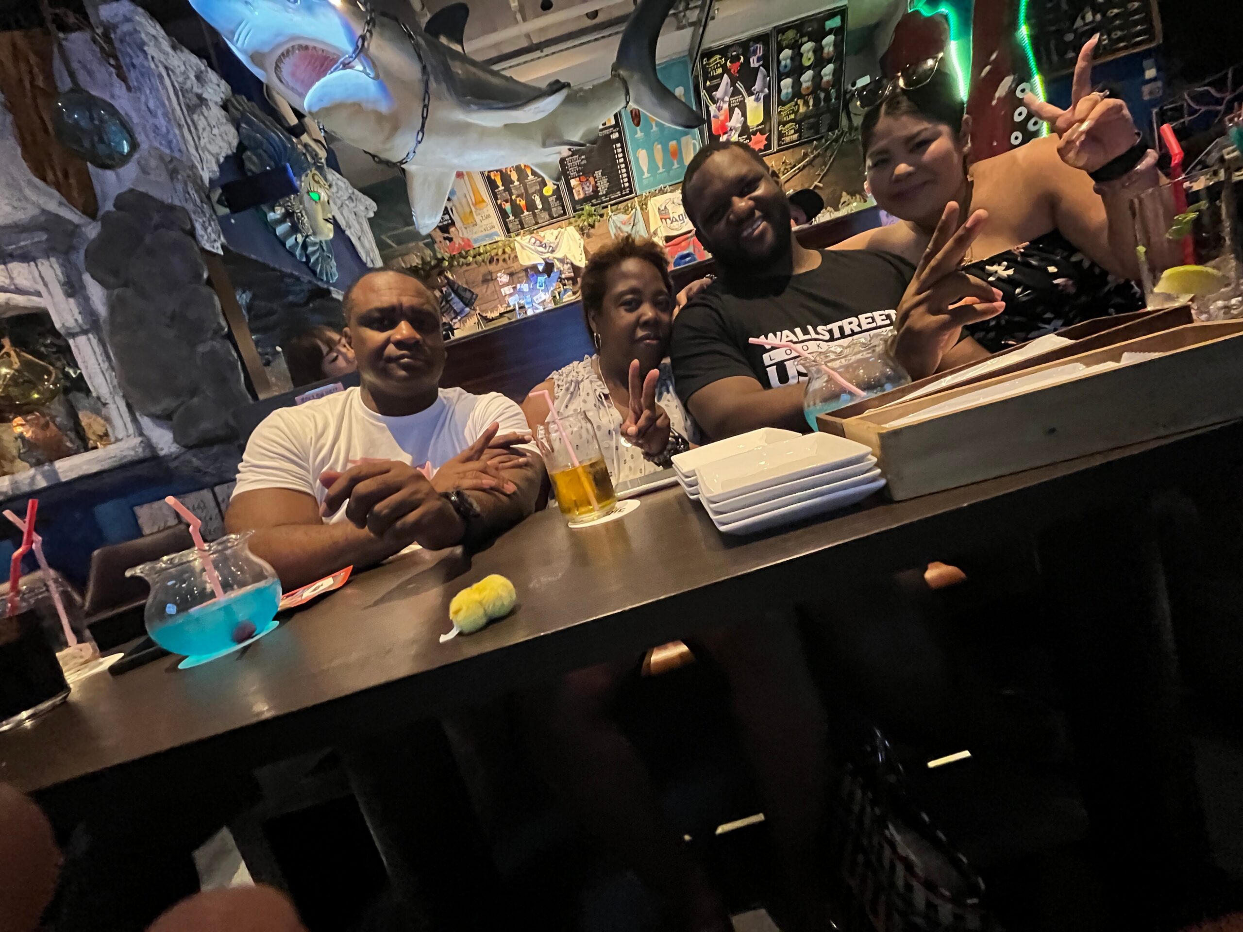 Group of five friends at a bar posing for a photo with a shark statue overhead and colorful drinks on the counter.