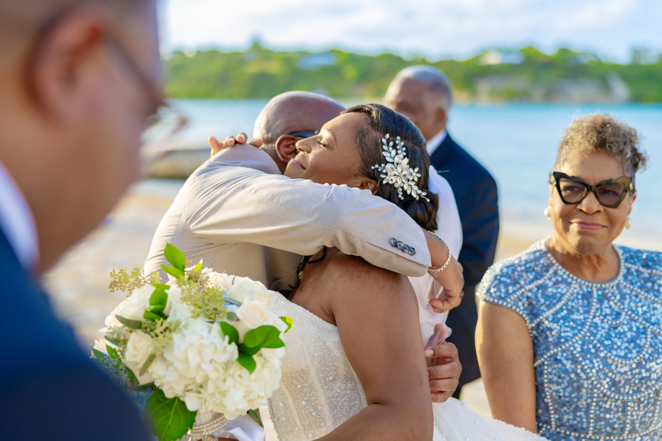 Bride in a white sequined dress hugs a man while holding a bouquet of white flowers by a waterfront.