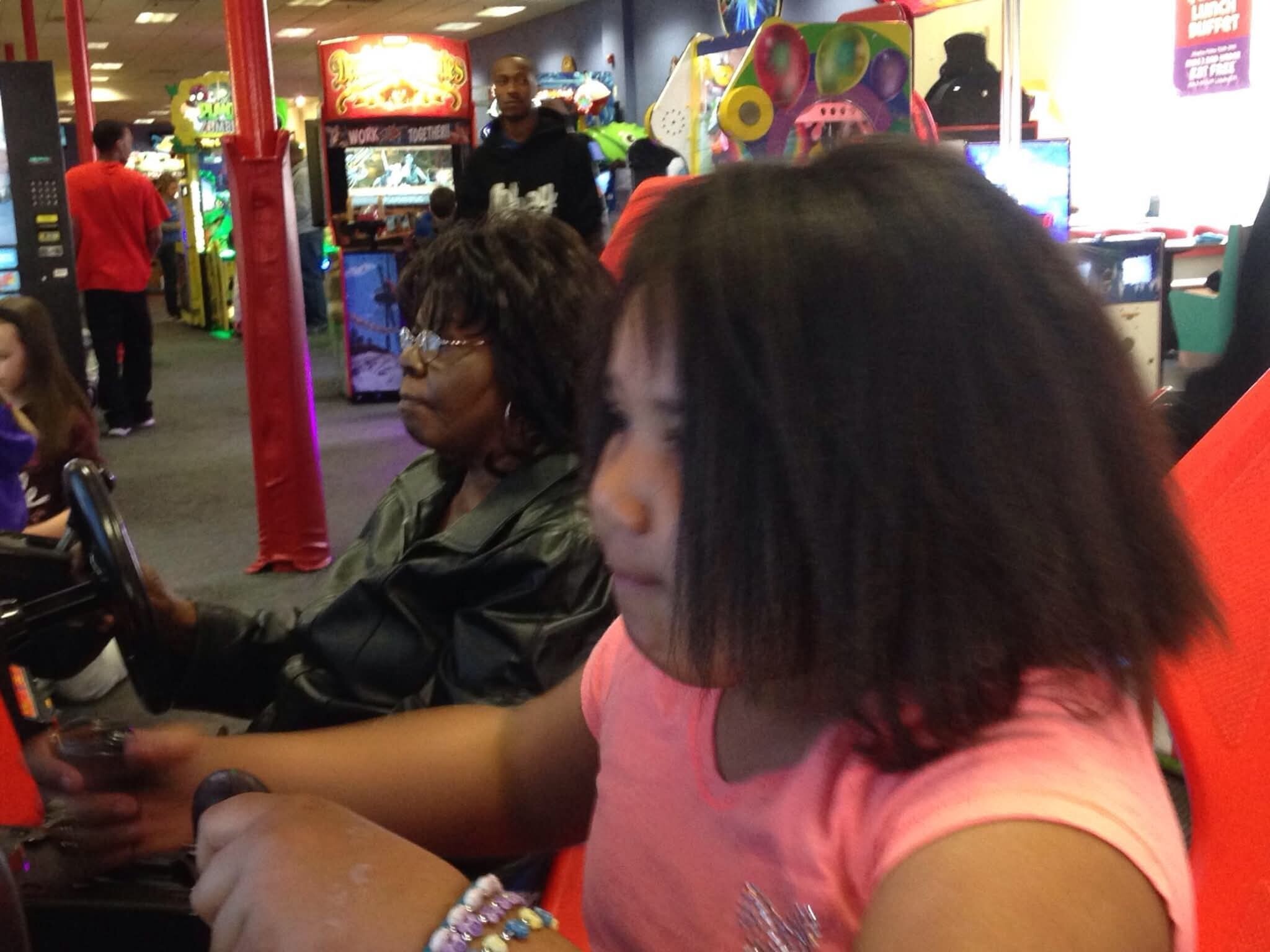 Foreground shows a child in a pink shirt playing an arcade game; colorful machines and a red padded pillar are in the busy arcade background.