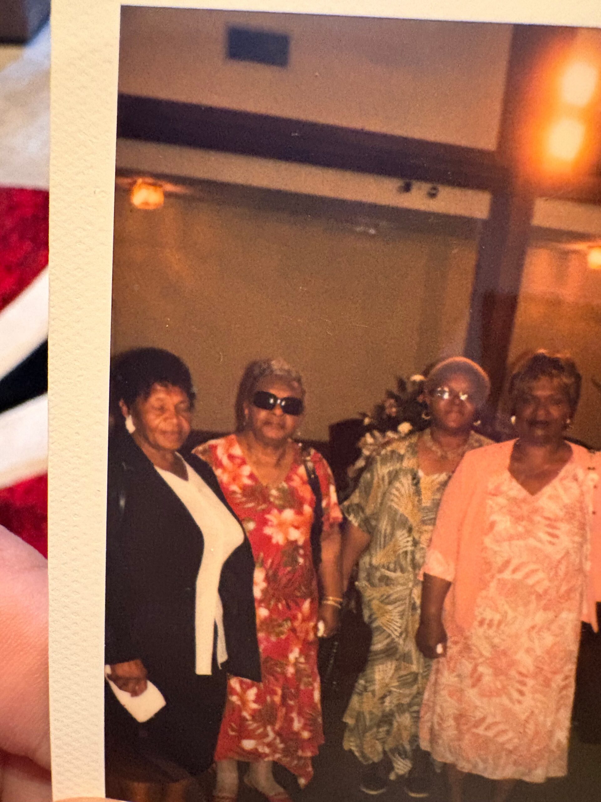 Group of four women posing together at a social event, wearing bright floral dresses and sunglasses in a warm indoor setting.