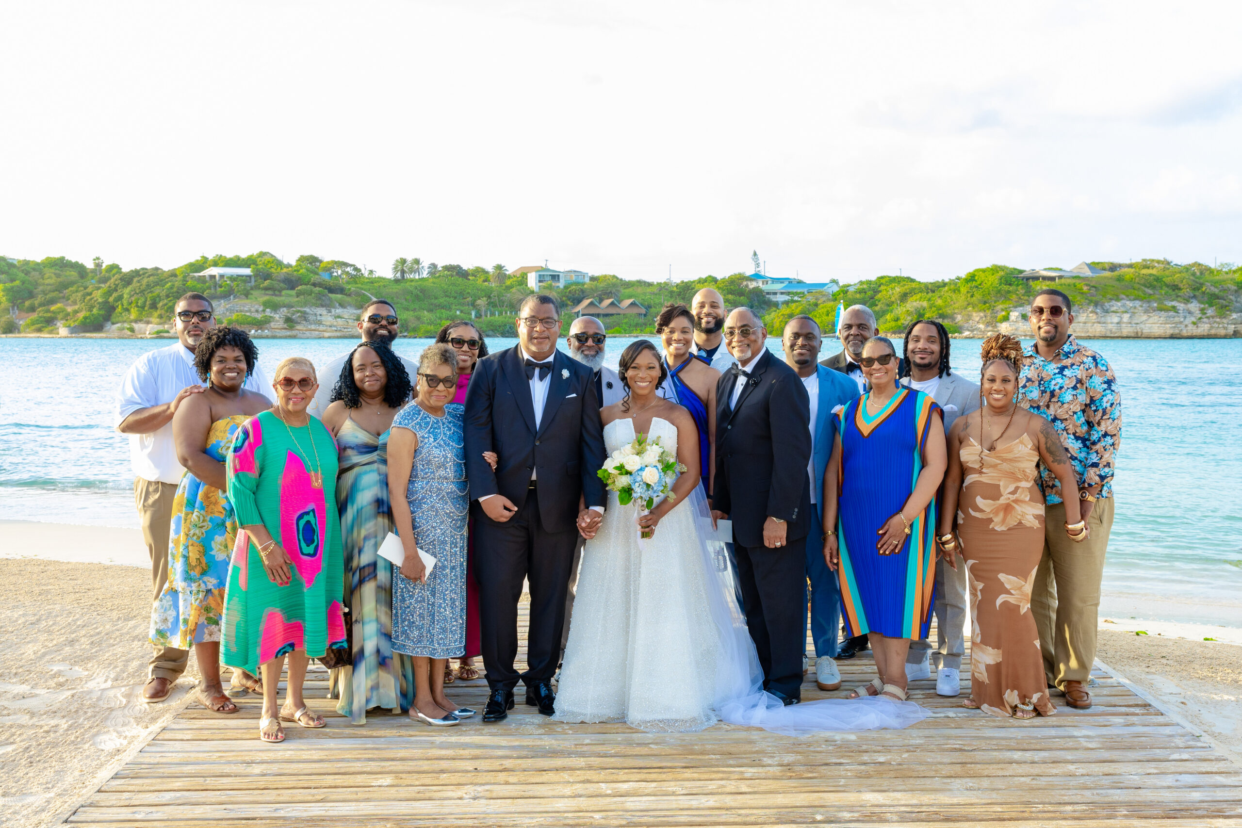 Wedding party group photo on a sunny beach with the bride and groom center, surrounded by family and friends in colorful outfits.
