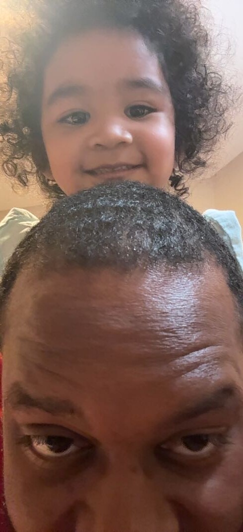 Child perched on an adult's shoulders, smiling at the camera; foreground shows the adult's forehead and eyes close up