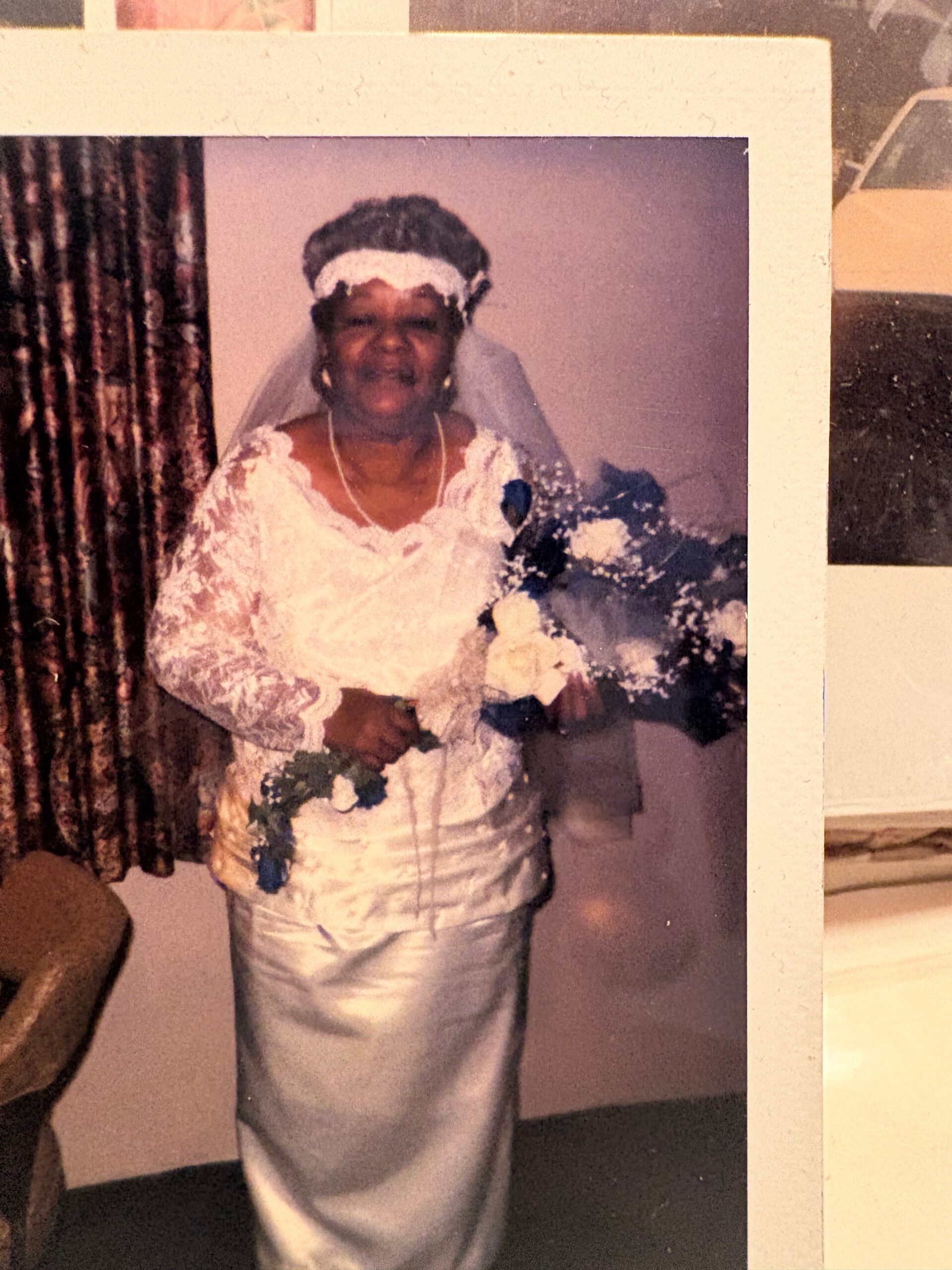 Older woman in a white wedding dress with veil and bouquet, standing indoors.