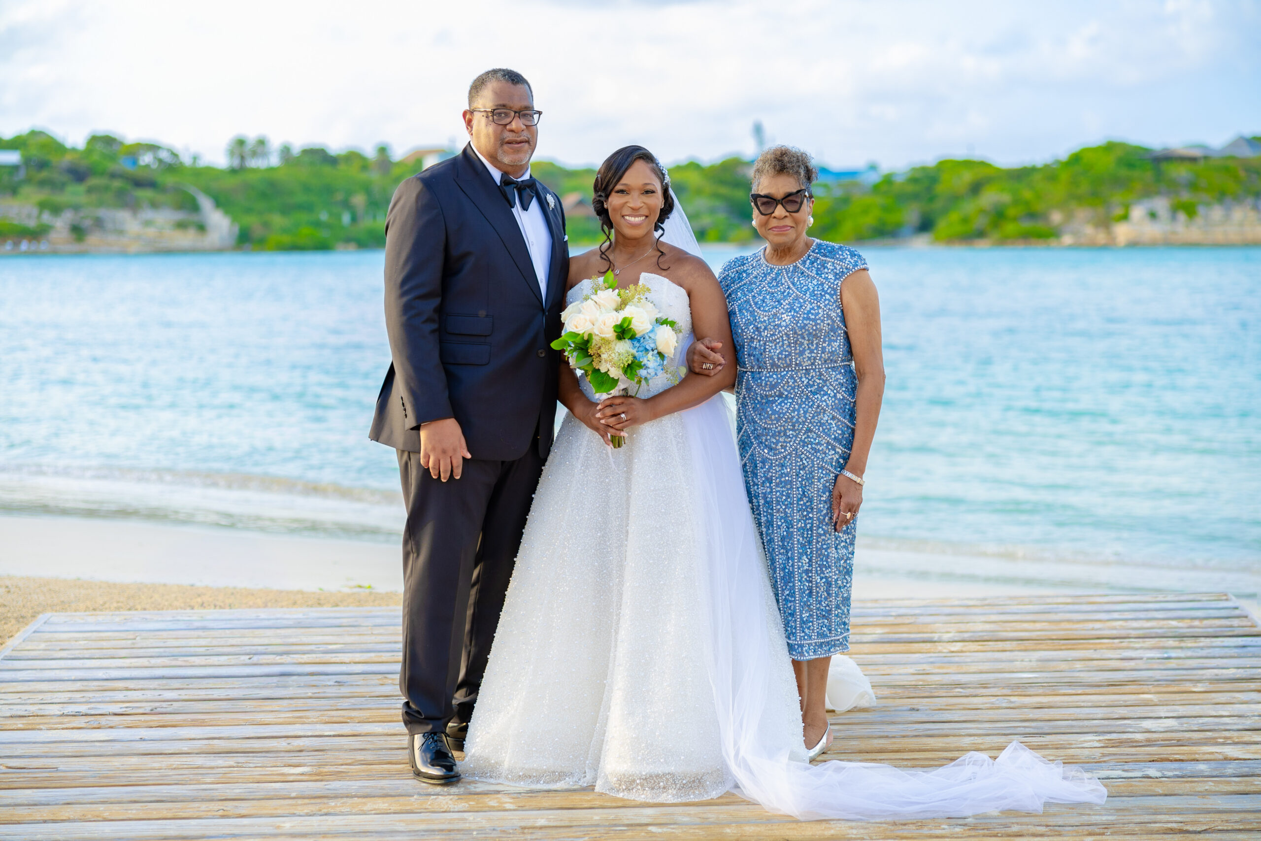 Bride in a sparkling white wedding dress holds a bouquet, flanked by a man in a black tuxedo and an older woman in a blue beaded dress, on a seaside boardwalk by calm water.