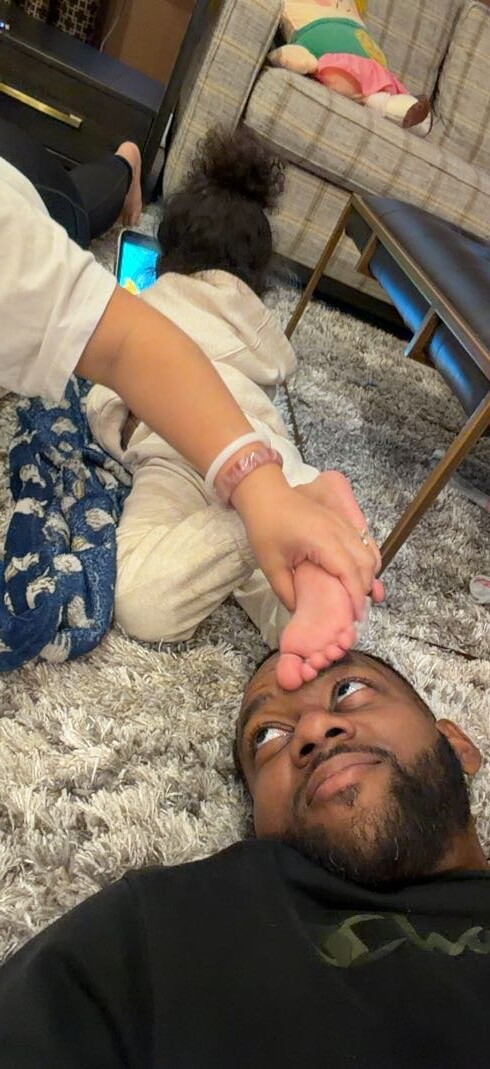 Man lying on a shaggy rug in a living room while a toddler playfully touches the man's toes near his mouth.