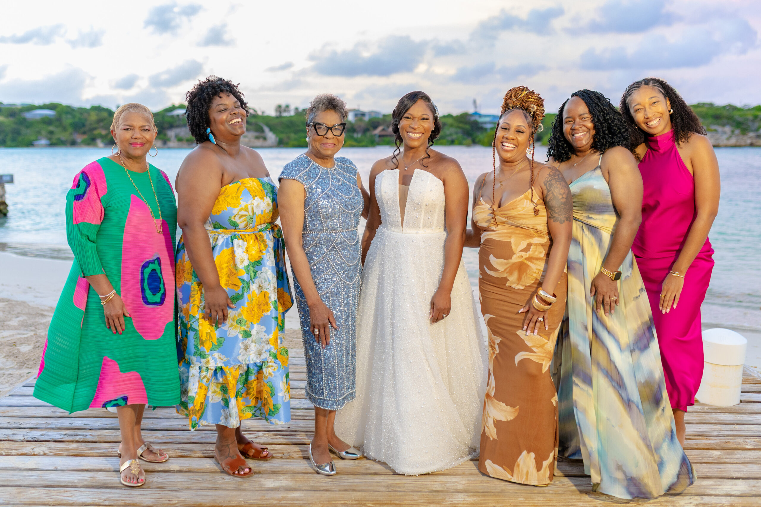 Group of eight women in bright dresses posing together on a wooden boardwalk by the seaside, smiling for a photo.