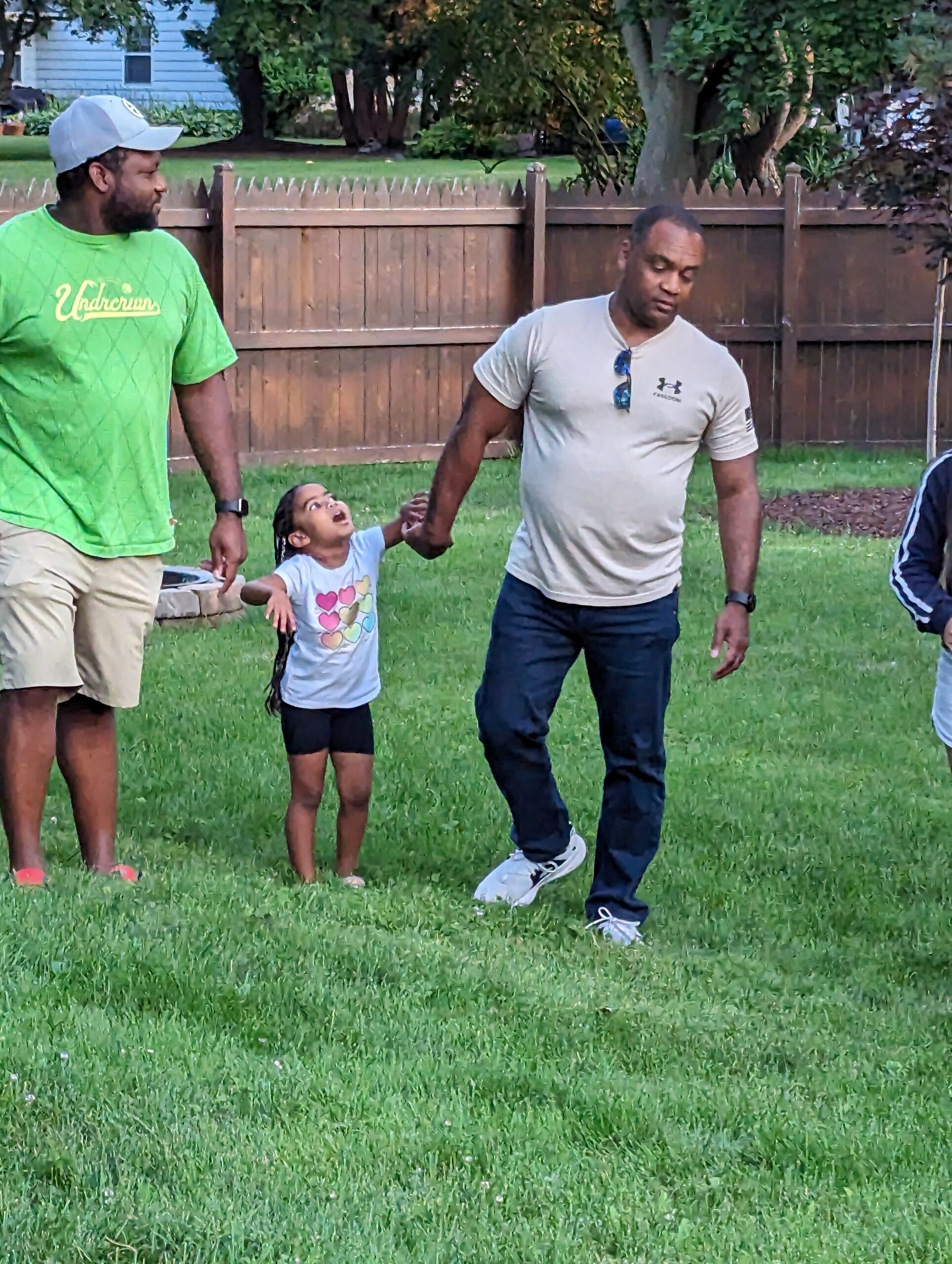 A child holds hands with two adults in a backyard as they walk across the grass, smiling and playing.