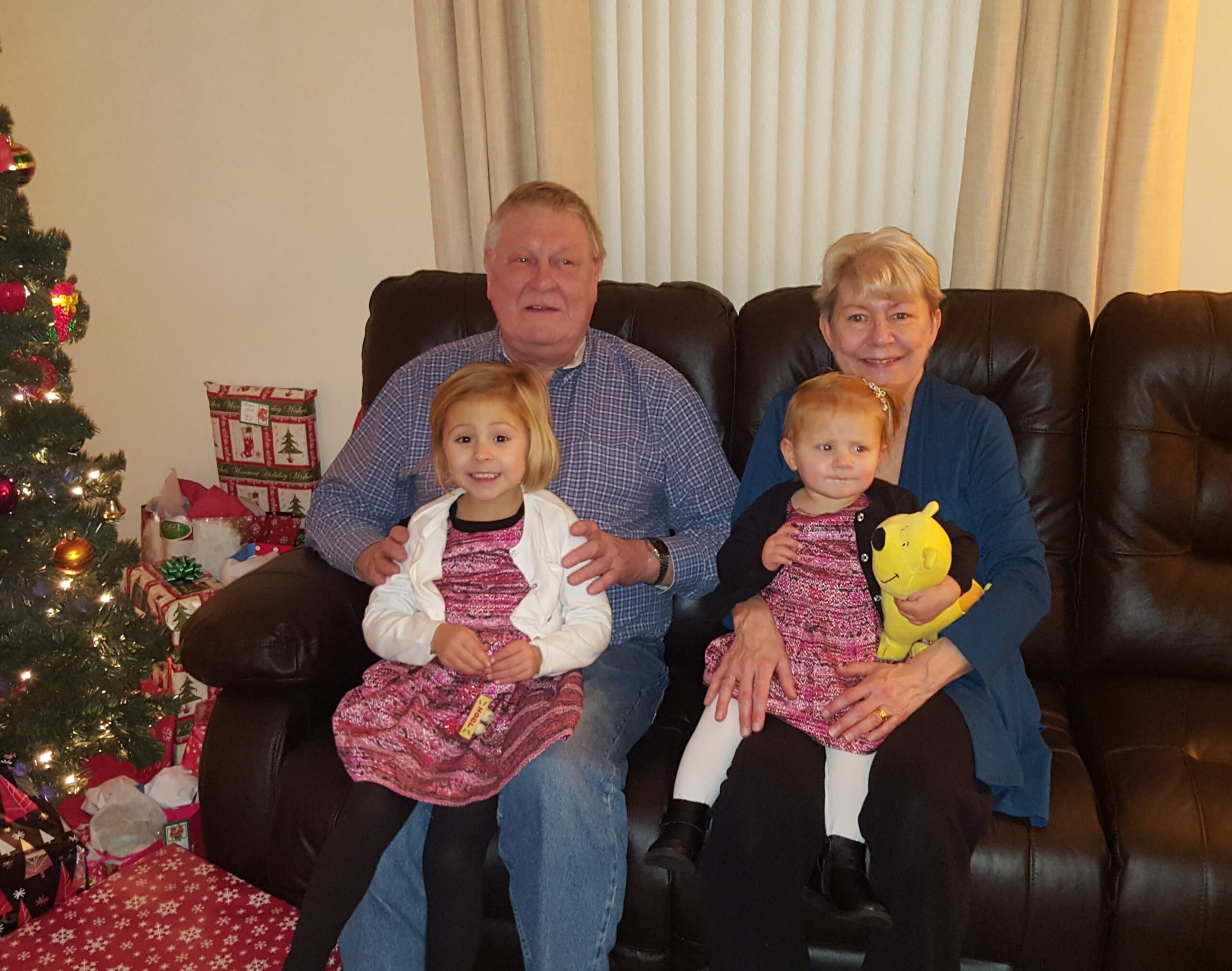 Family seated on a couch with two young girls, Christmas tree and gifts nearby, celebrating the holidays.