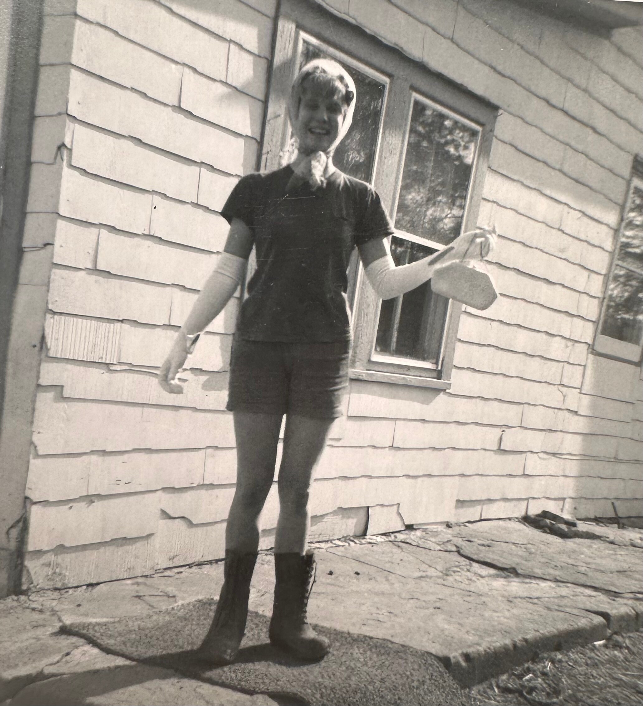 Black-and-white photo of a person standing on a sidewalk in front of a clapboard house, wearing a dark dress and light arm wraps, smiling.