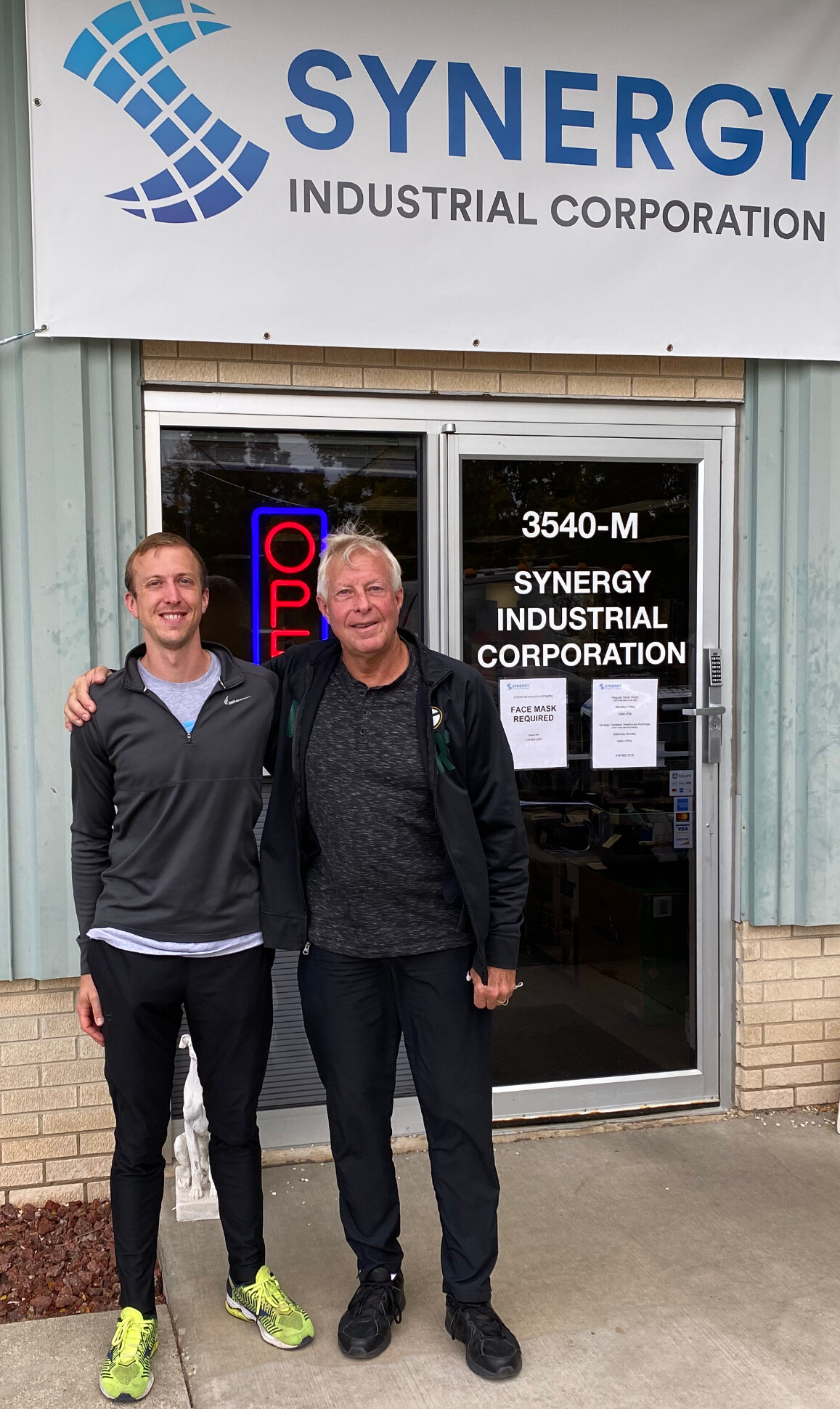 Two men standing outside Synergy Industrial Corporation, smiling, in front of the entrance sign reading 3540-M.