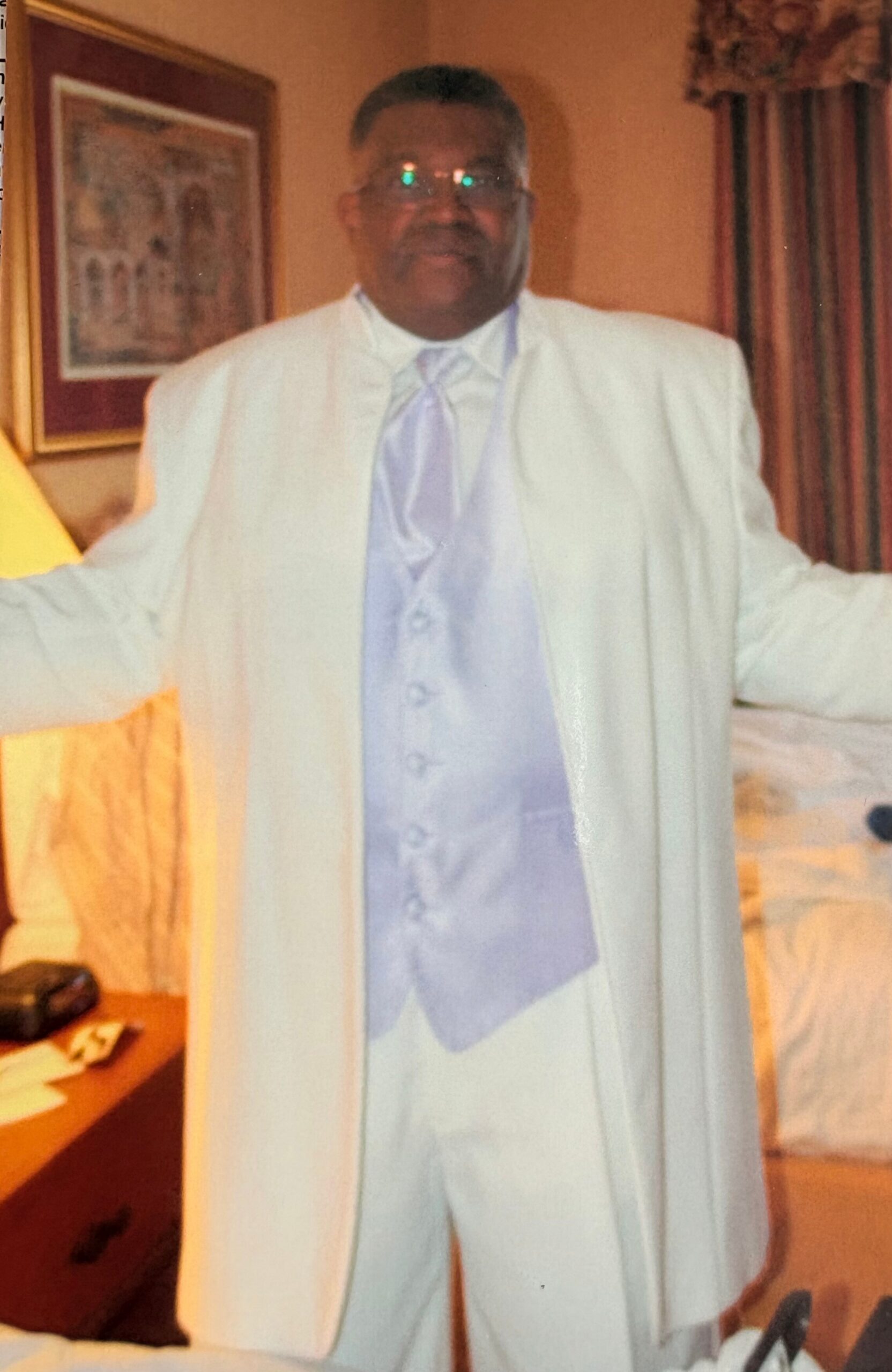 Man in a white suit with a light blue vest and tie, standing indoors with arms outstretched.