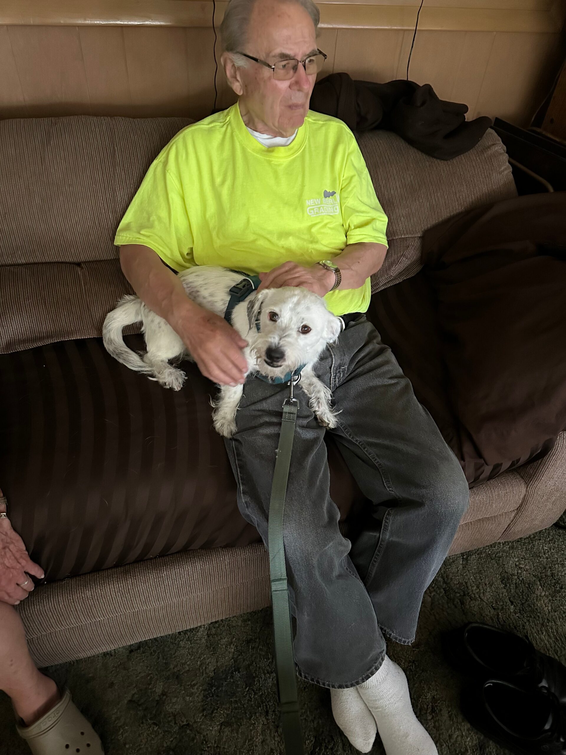 Elderly man in a neon yellow shirt sits on a brown couch with a small white dog on his lap, wearing a blue collar and leash.