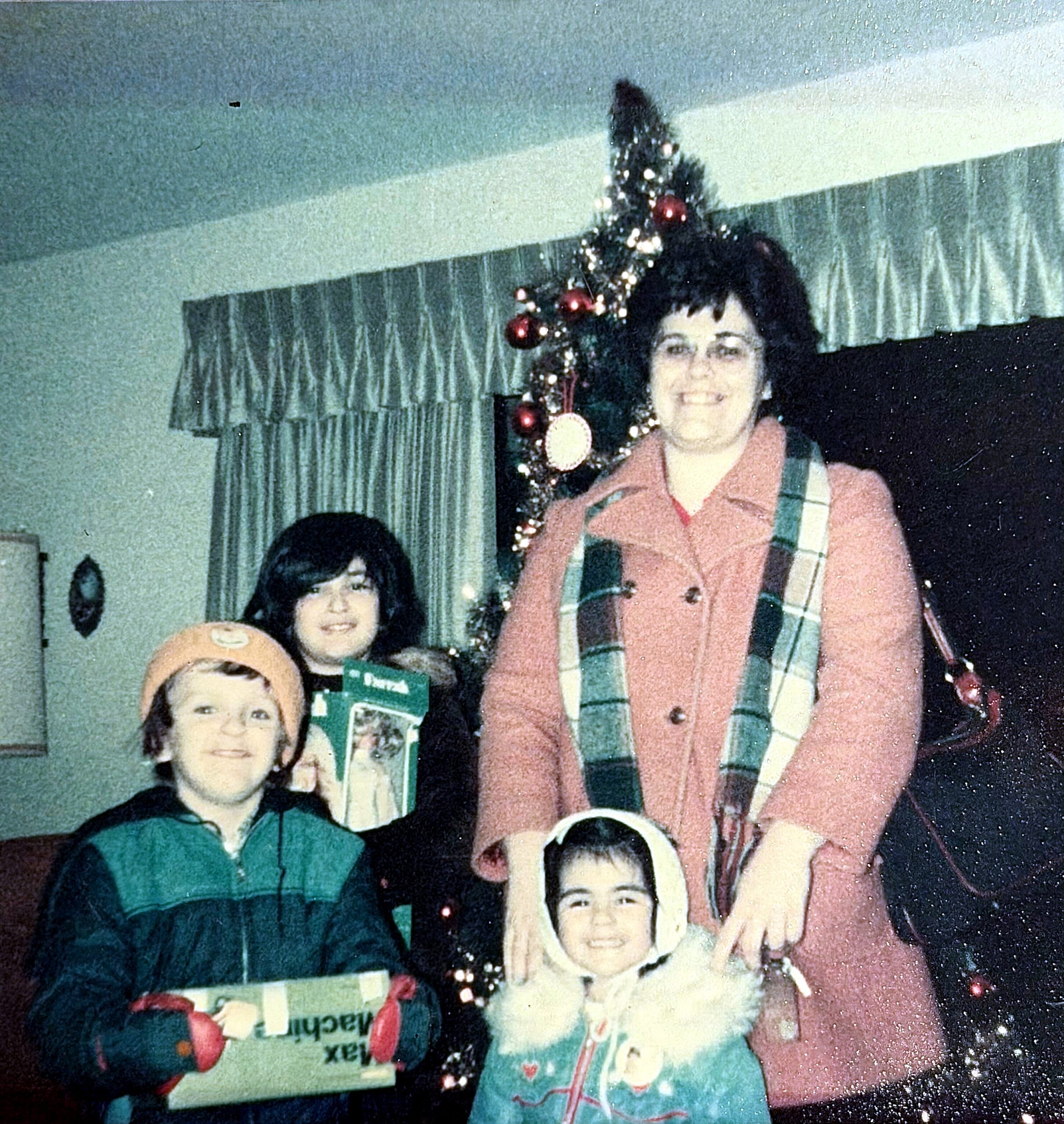 Woman in pink sweater standing beside a decorated Christmas tree, with children and gifts in a living room.