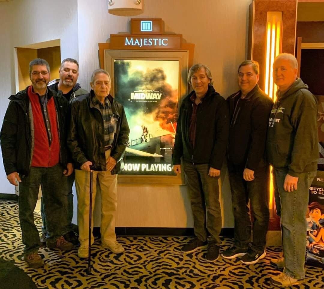 Six men in jackets pose together in a cinema lobby in front of a Midway poster at the Majestic theater sign above them.