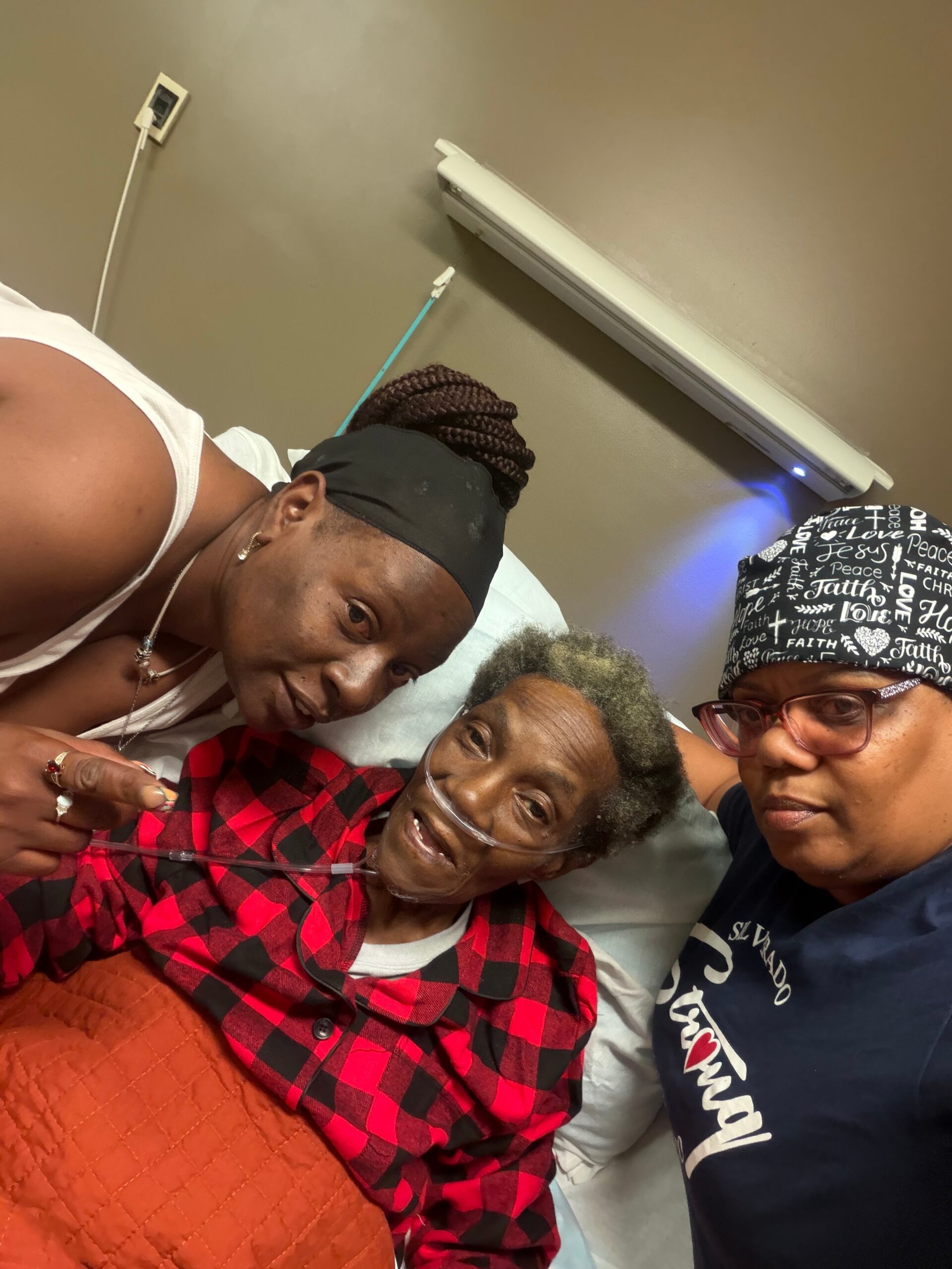 Three women sit closely together in a hospital room, one patient wearing an oxygen mask and red plaid shirt, flanked by two visitors.
