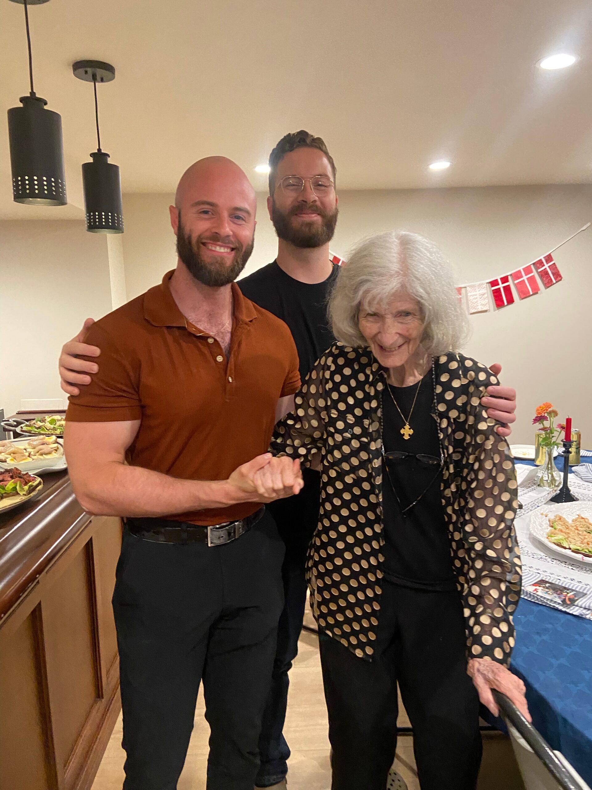 Smiling trio at a party: two men and an elderly woman in a polka-dot jacket posing by a buffet with Danish flag bunting in the background.