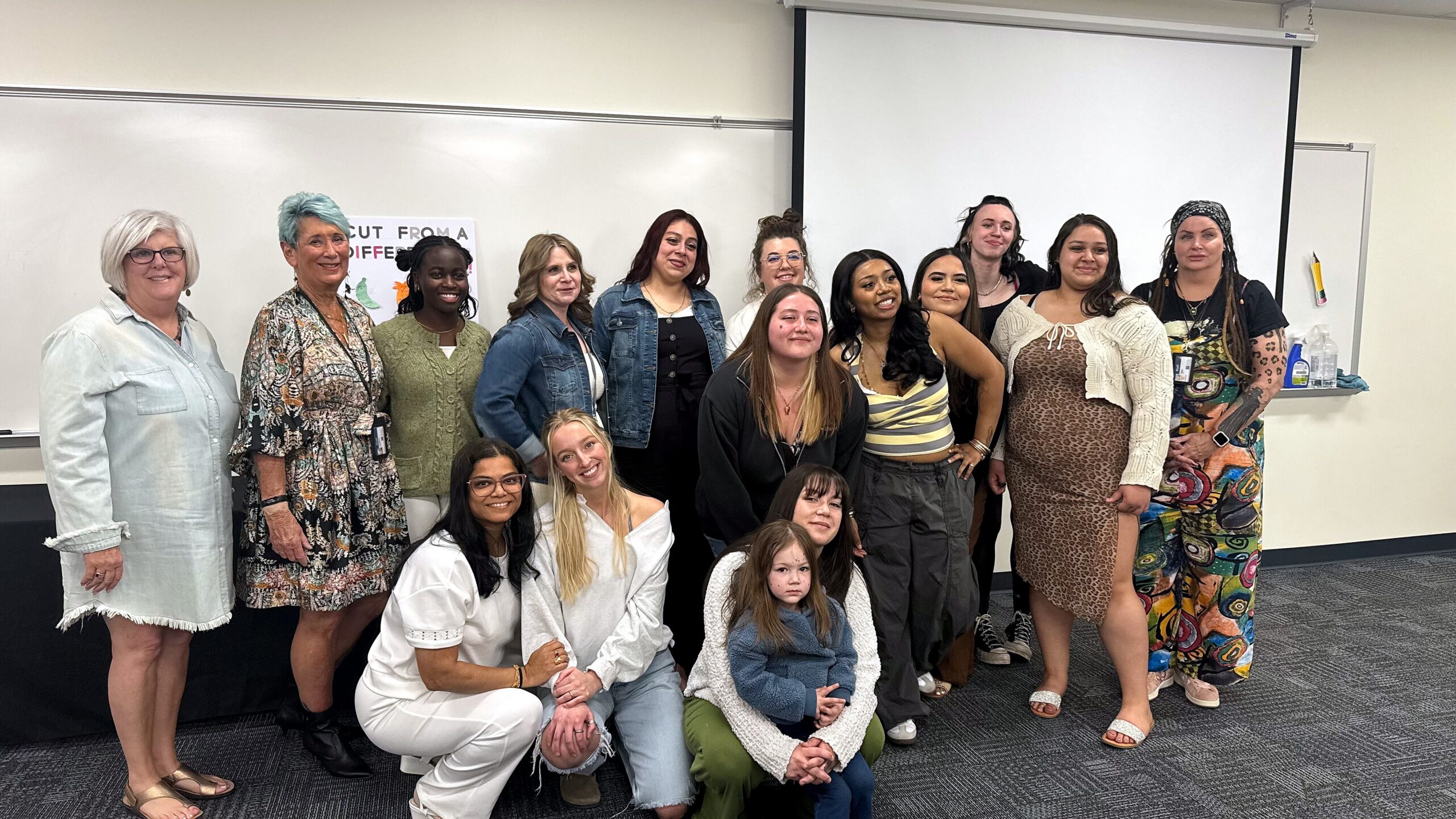 Group of diverse women (and a few kids) posing together in a classroom for a group photo, with a whiteboard behind them and a projector screen above head height.