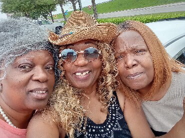 Three women posing for a selfie outdoors, smiling at the camera; one wears a straw hat and sunglasses.