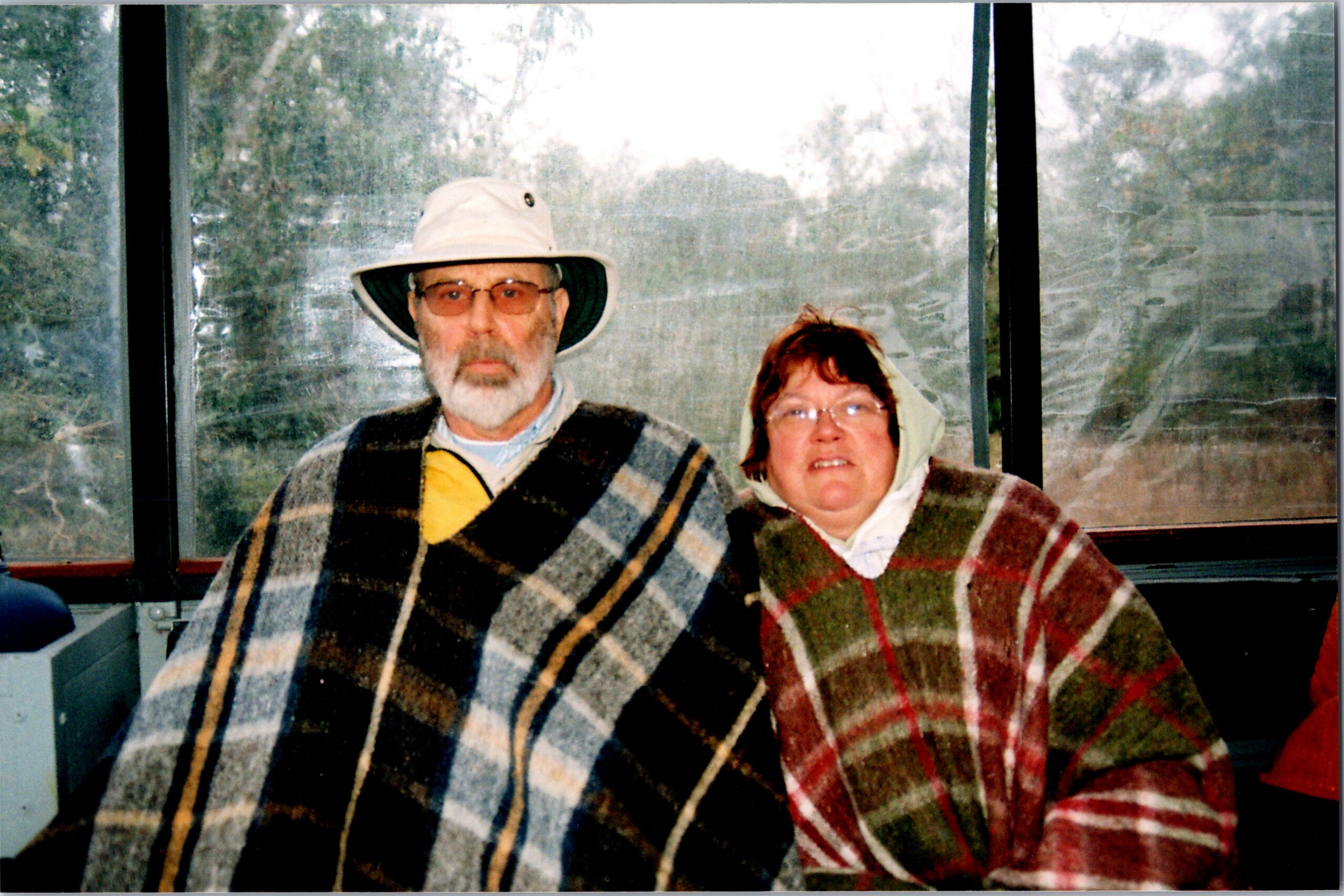 Two elderly people sit closely together, each wrapped in a large plaid blanket, and look toward the camera inside a vehicle.