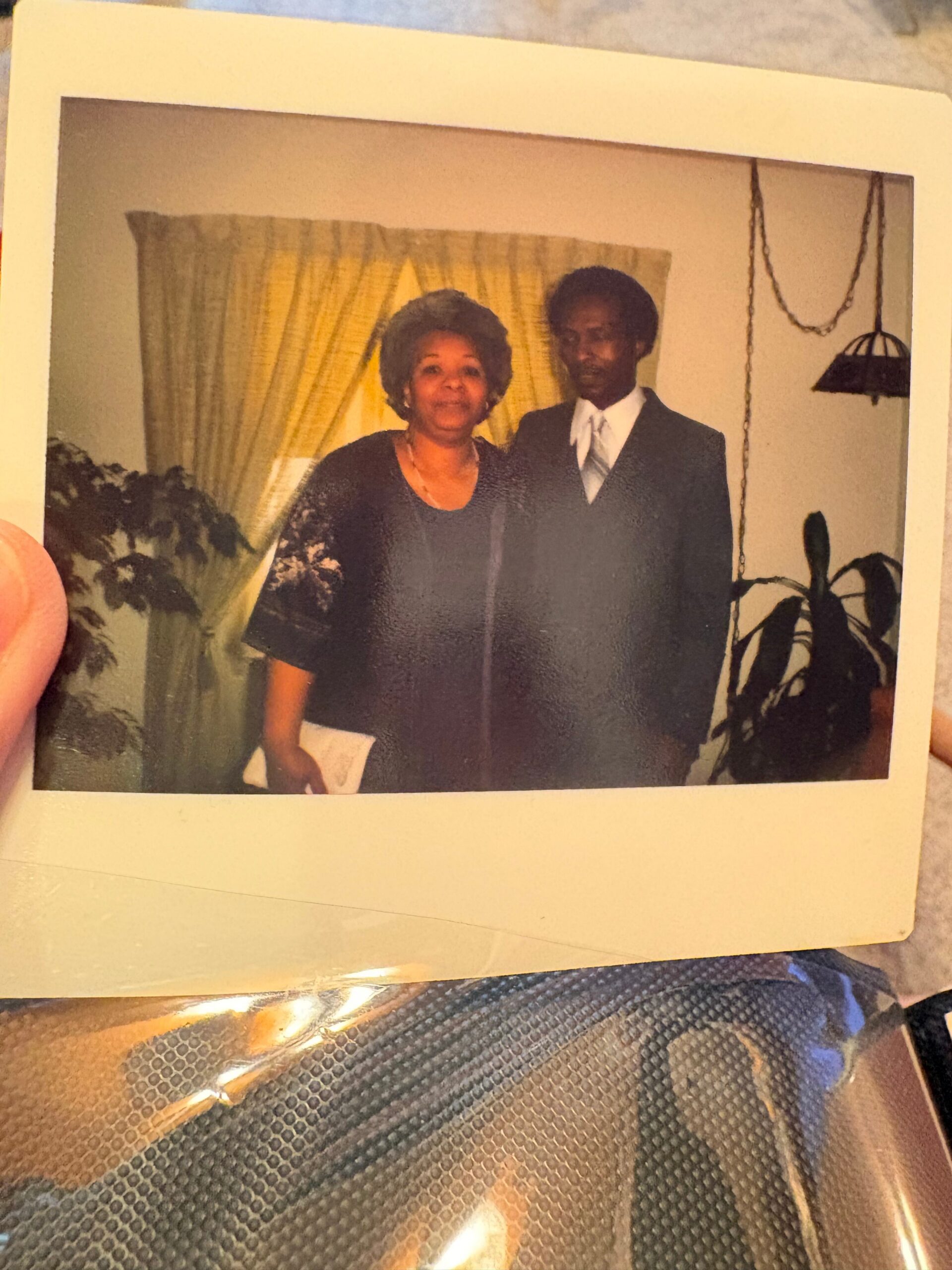 Two people stand together indoors: a smiling woman in a dark dress on the left and a man in a suit on the right, with yellow curtains behind them.