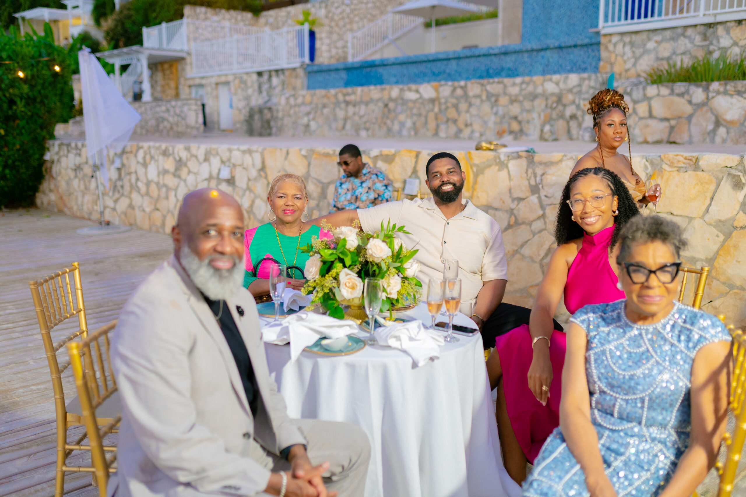 Group of adults at an outdoor celebration seated around a white-clothed table with a flower centerpiece, smiling at the camera. Stone wall and blue tiled backdrop in the background.