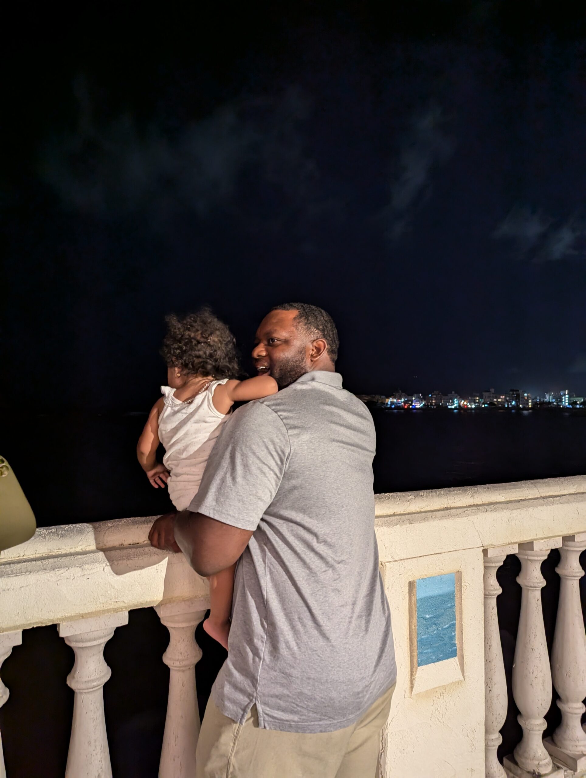 Man holding a child while leaning on a seaside railing at night, city lights in the distance behind them.