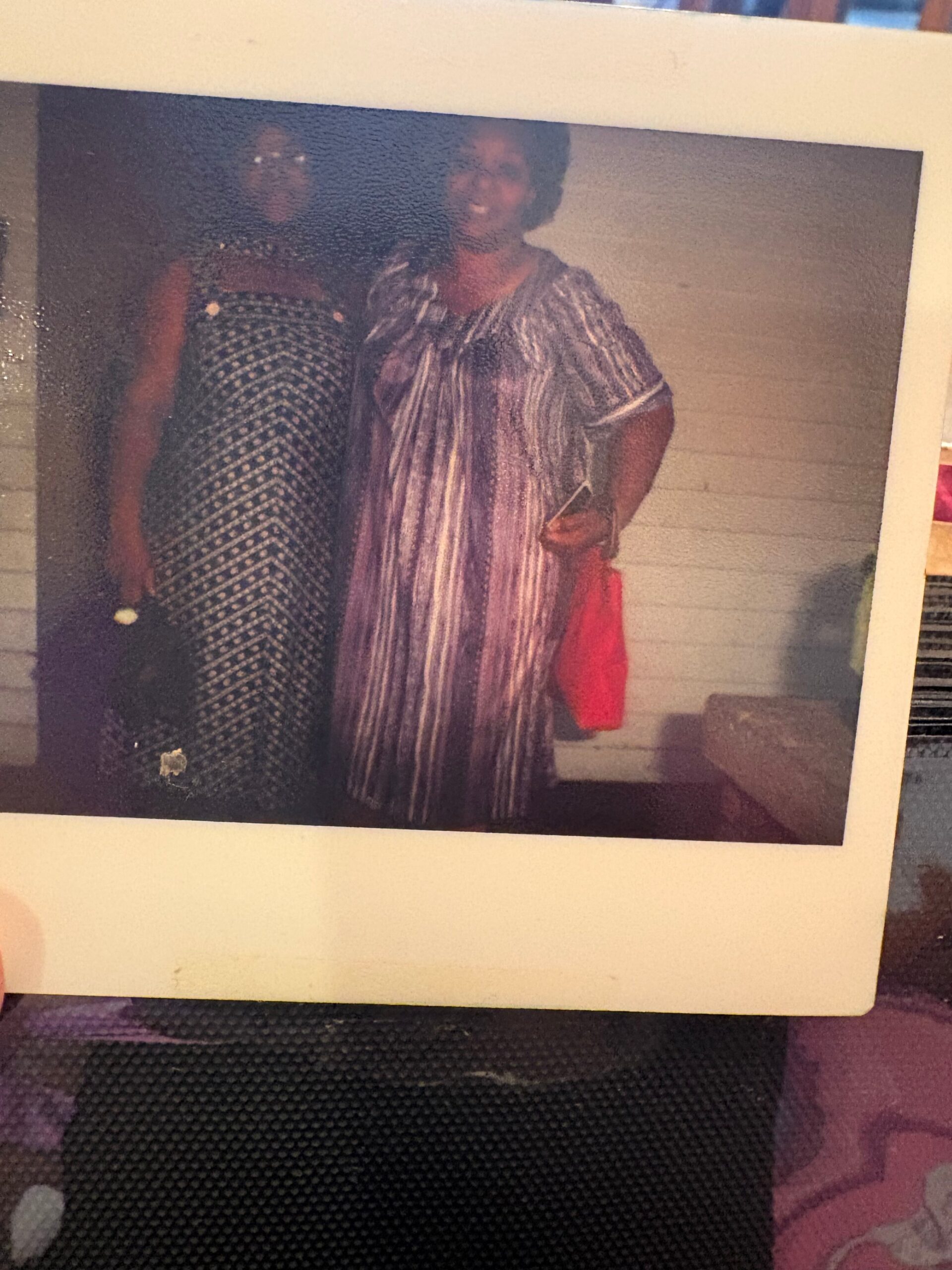 Two women posing together indoors, smiling at the camera; one wears a black-and-white patterned dress, the other in a purple striped dress holding a red bag and a phone.