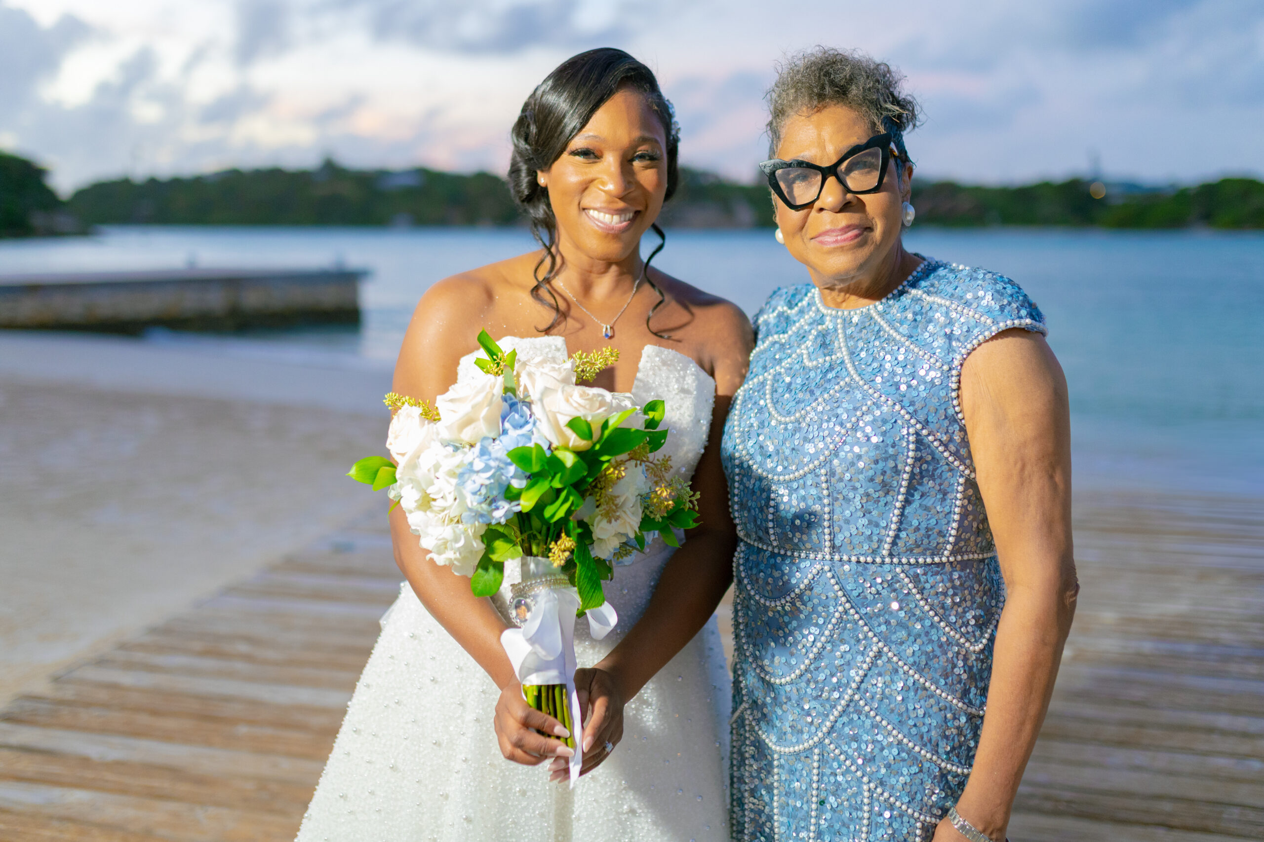 Bride in a white wedding gown holding a bouquet, standing with an older woman in a blue sequined dress on a beach.