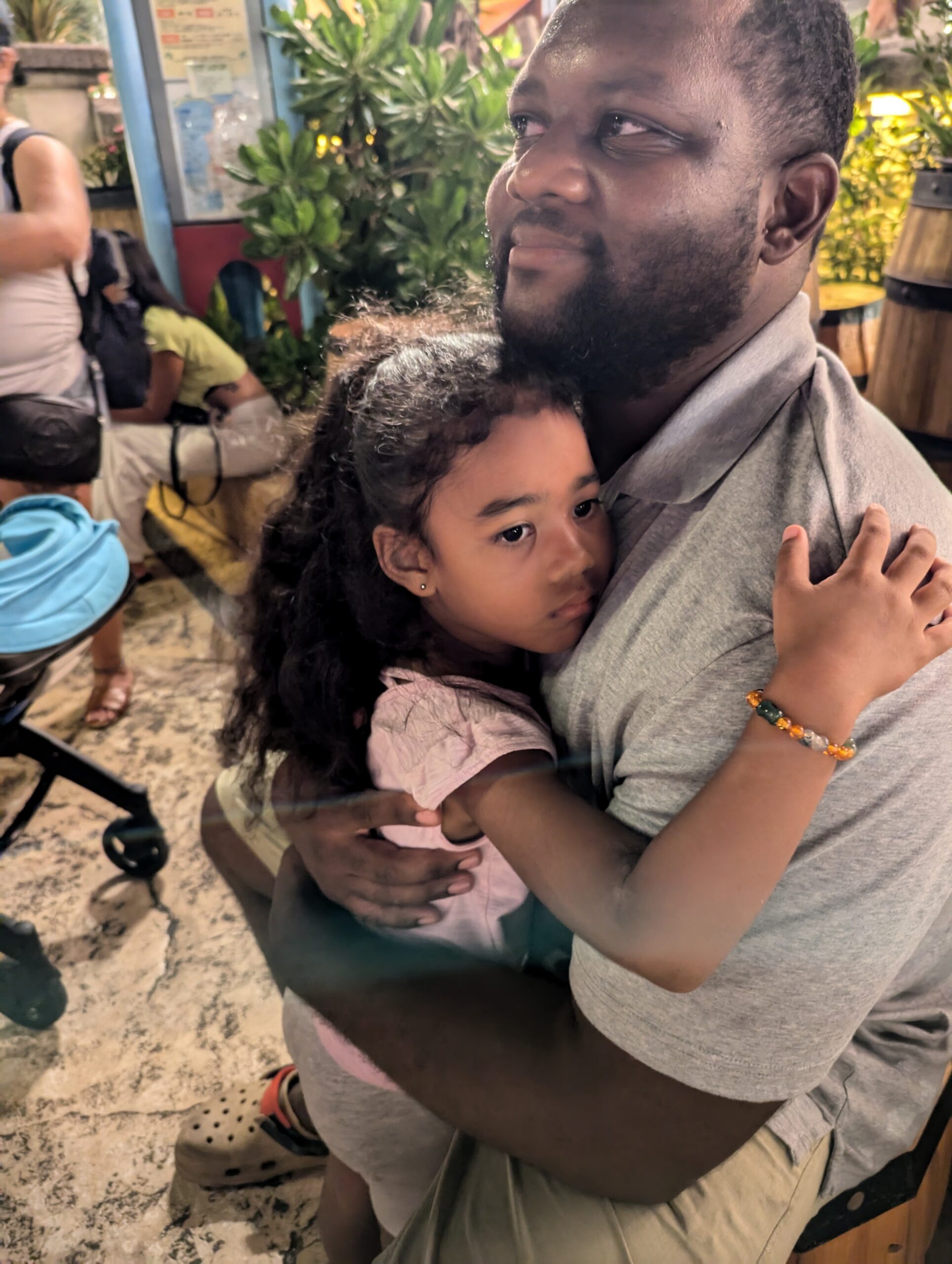 A man in a gray shirt hugs a young girl in a pink top, both looking off to the side in a cozy, plant-filled setting.
