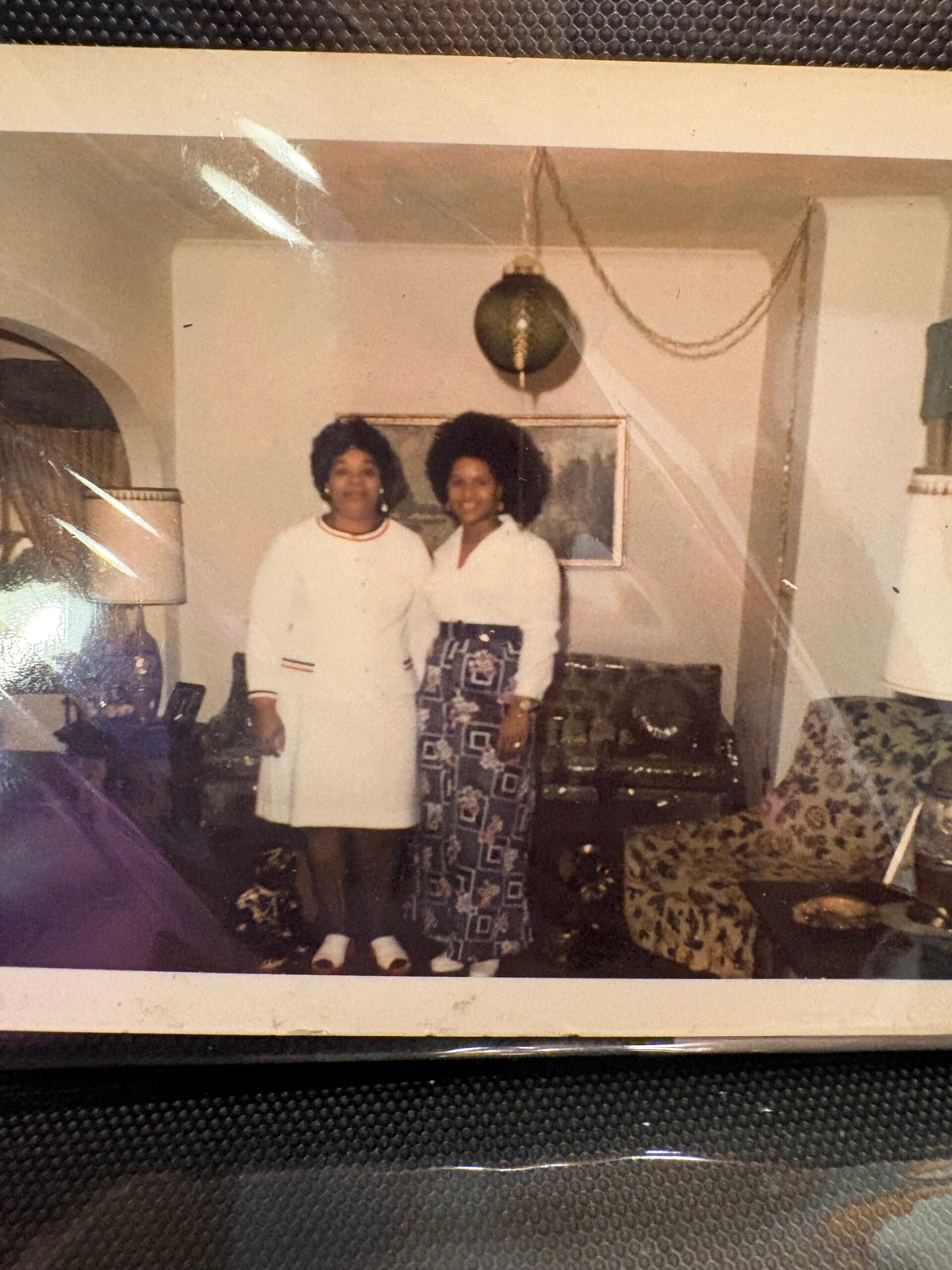 Two women stand together in a cozy living room, posing for a photo; one wears a white dress and the other a patterned skirt with a white blouse.