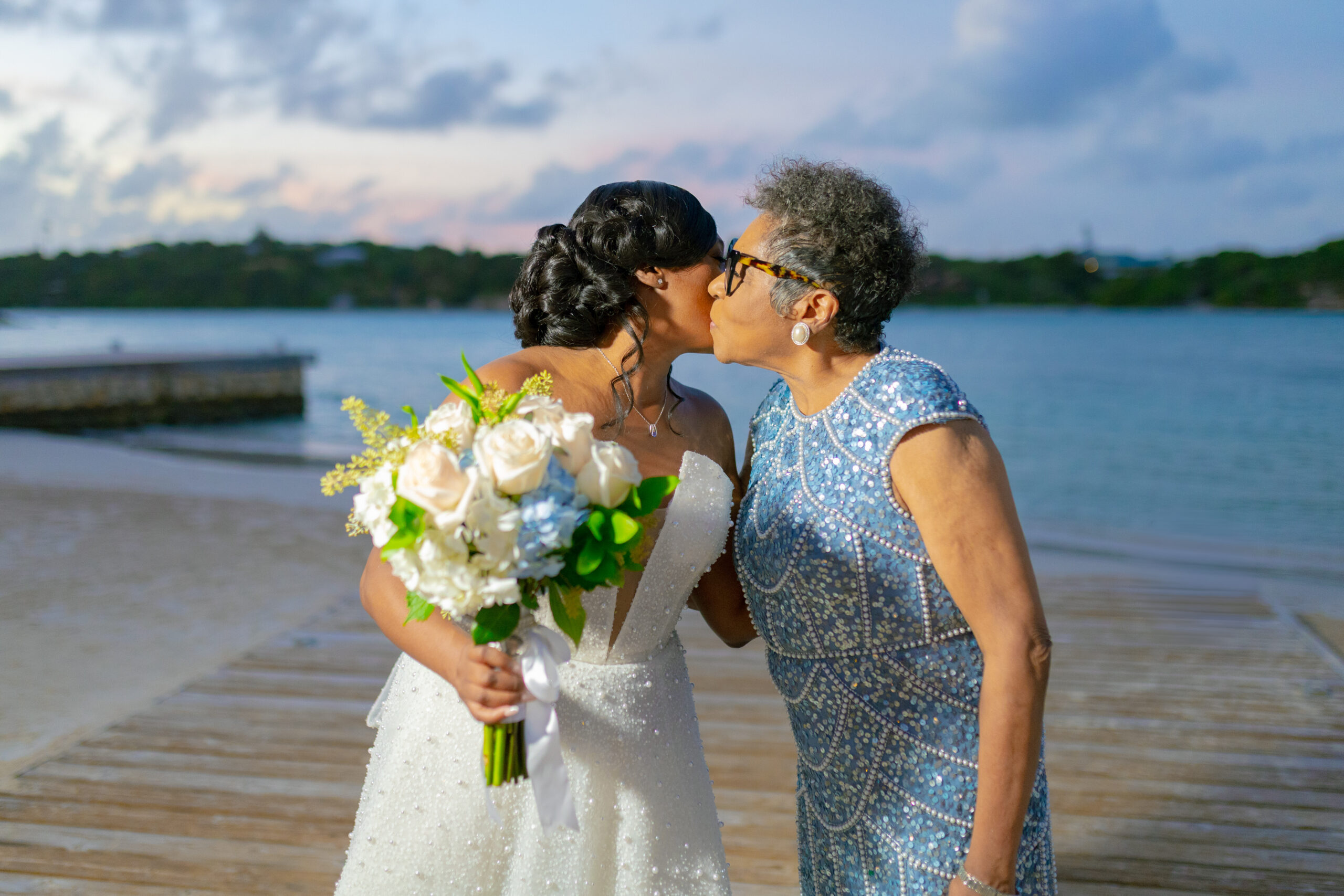 Two women kiss on a sandy beach; one in a white wedding dress holding a bouquet, the other in a blue sequined dress nearby.
