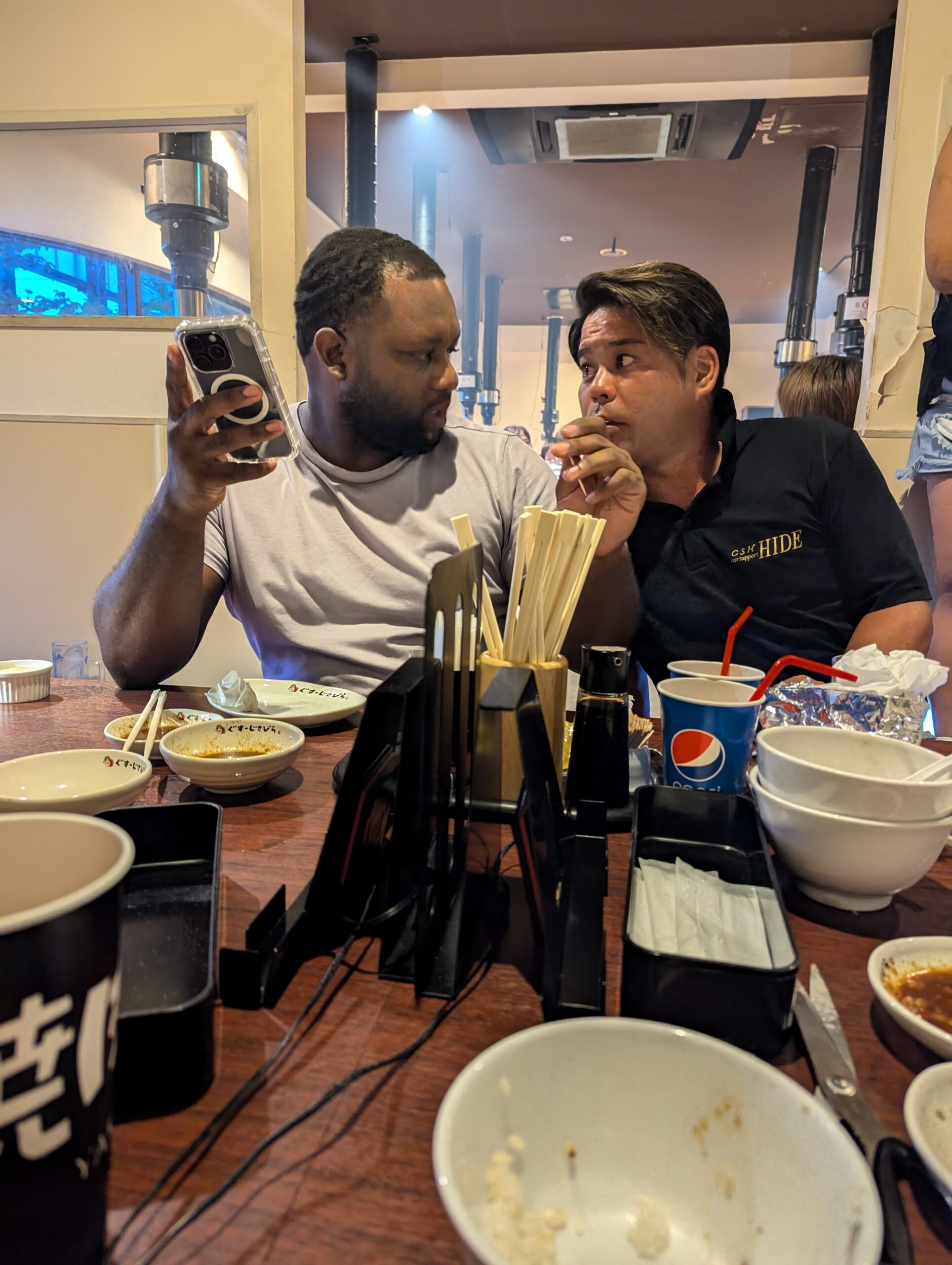 Two men sit at a restaurant table with bowls of food and chopsticks; one man is looking at his phone while the other talks, as drinks and condiments clutter the table.