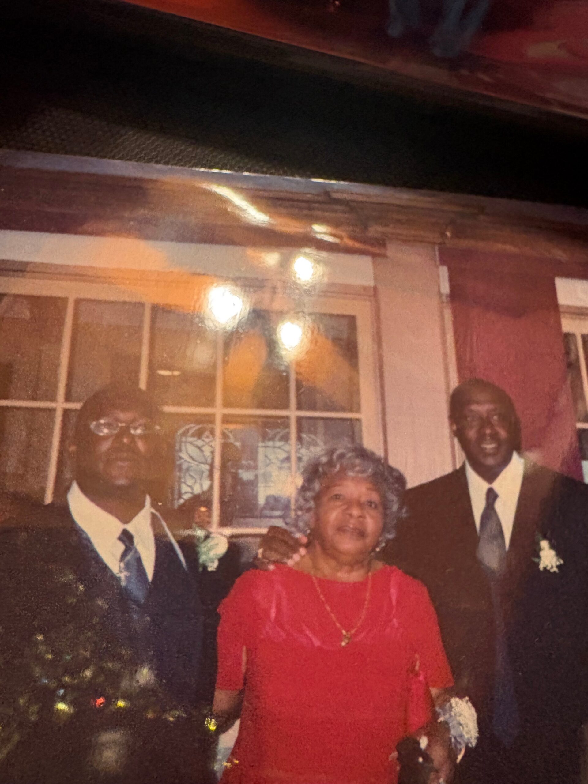 Older woman in a red dress standing with two men in suits at a nighttime indoor event, smiling for the photo.