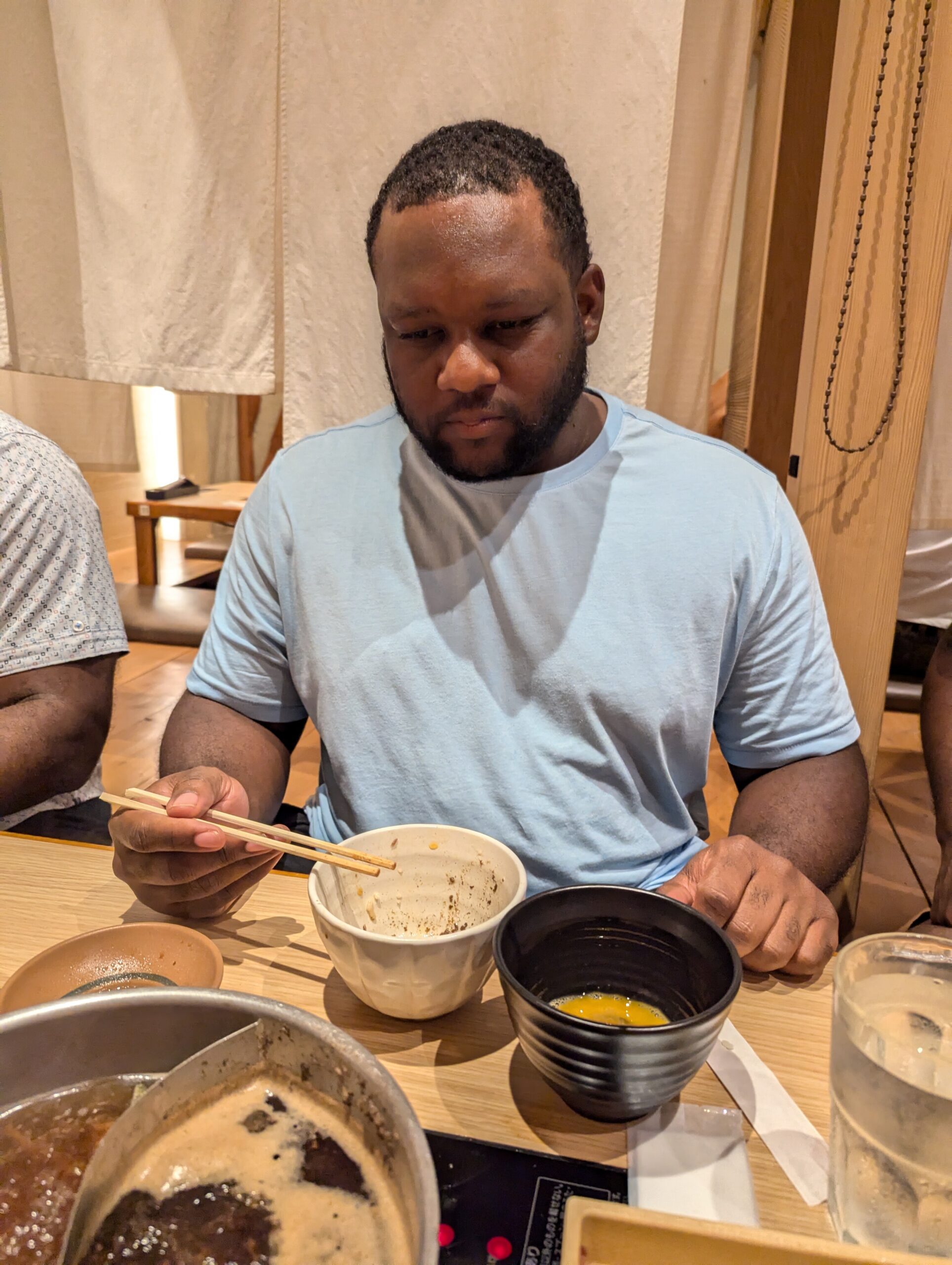 Man in a light blue shirt eating with chopsticks at a wooden table, bowls in front of him.