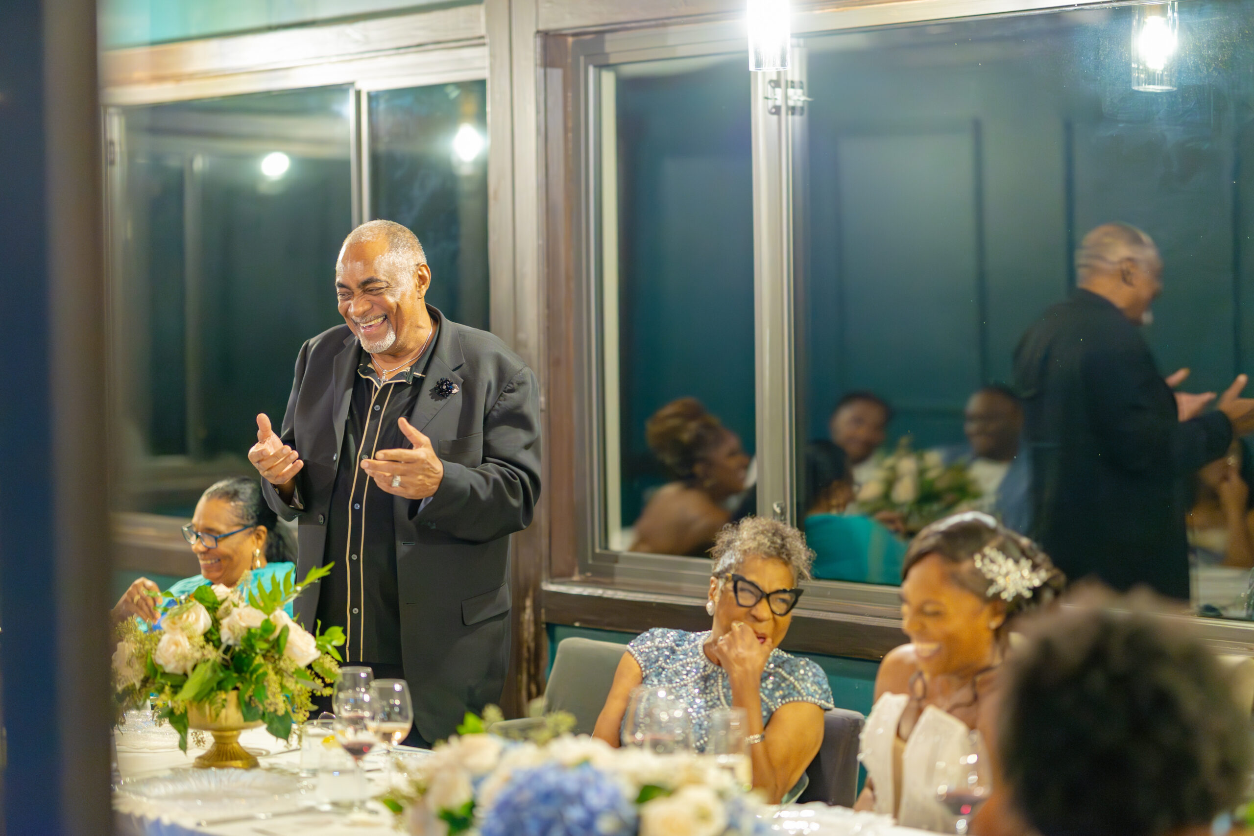 Smiling man in a black suit speaking at a banquet table with guests and a floral centerpiece nearby.