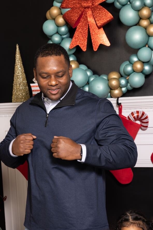 Man flexing his fists in front of a Christmas wreath and stockings, smiling in a navy pullover.