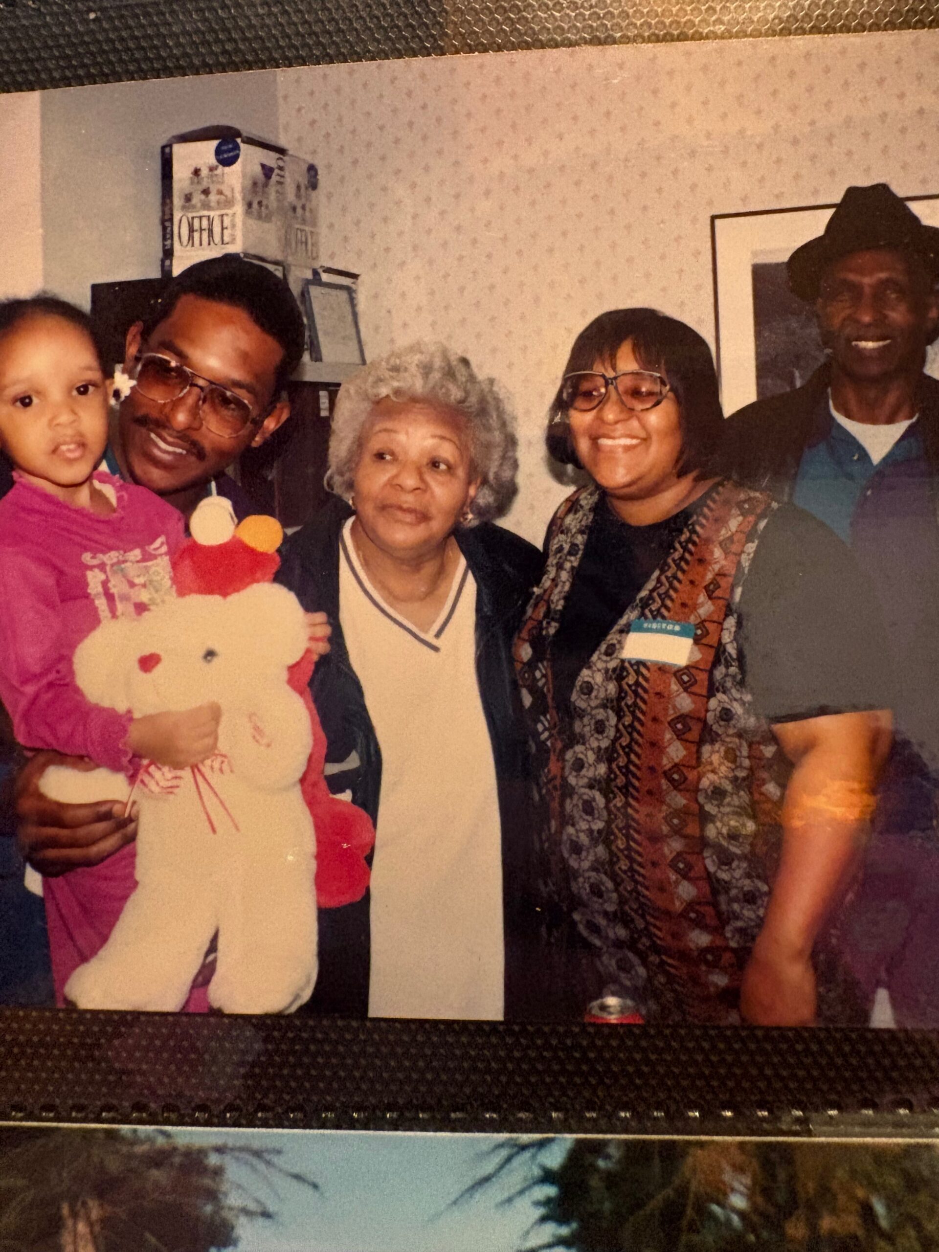 A multi-generational family posing together indoors, with a child holding a teddy bear and four adults smiling.
