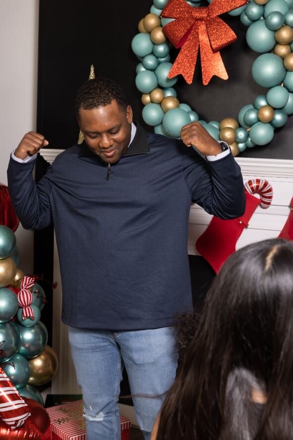 Man flexing his arms at a Christmas party, standing before a teal and gold balloon wreath with a red bow and stockings behind him.
