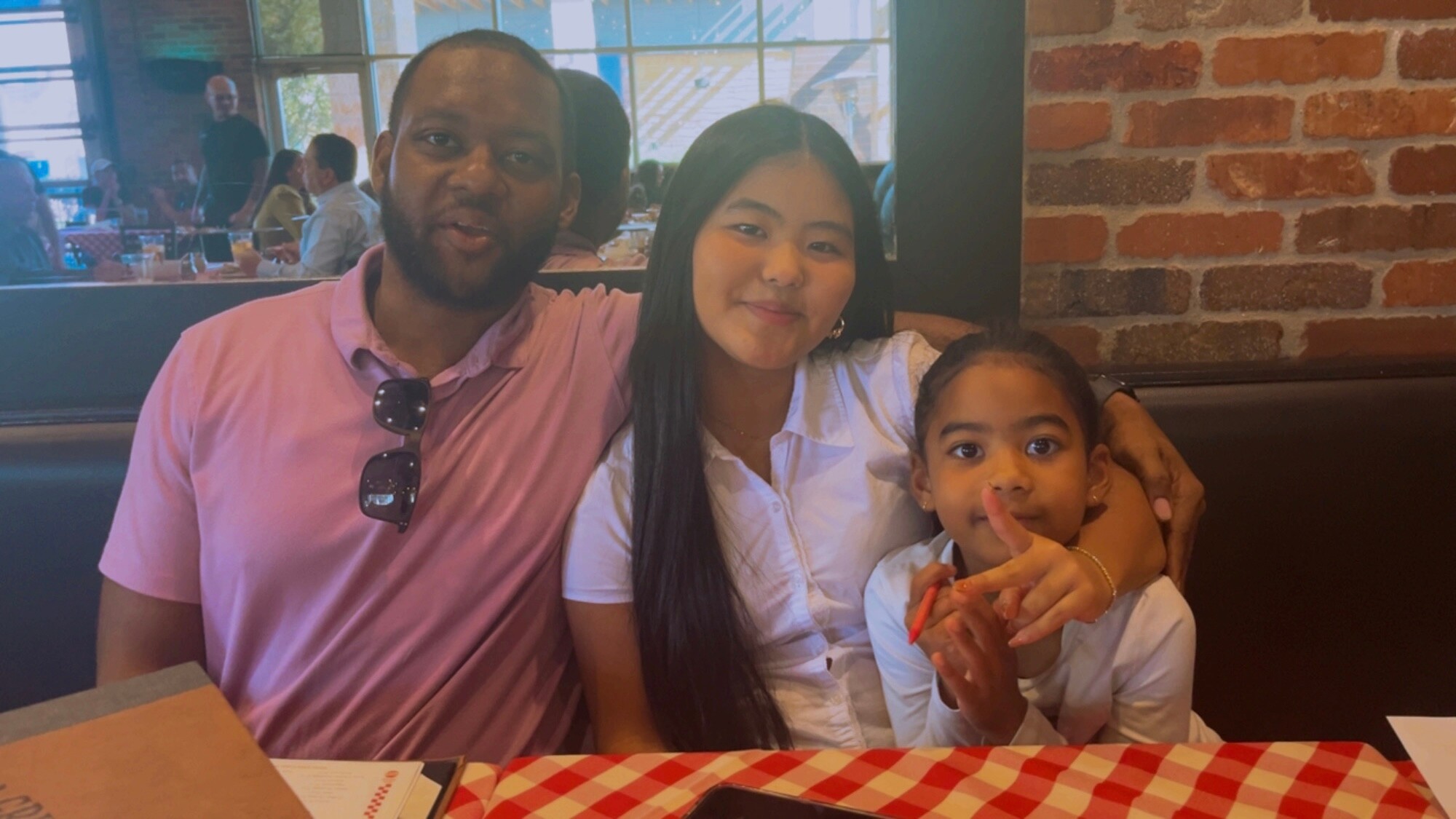 Family portrait in a restaurant booth: man in pink shirt with sunglasses, woman with long black hair, and a young girl in white, all smiling together.