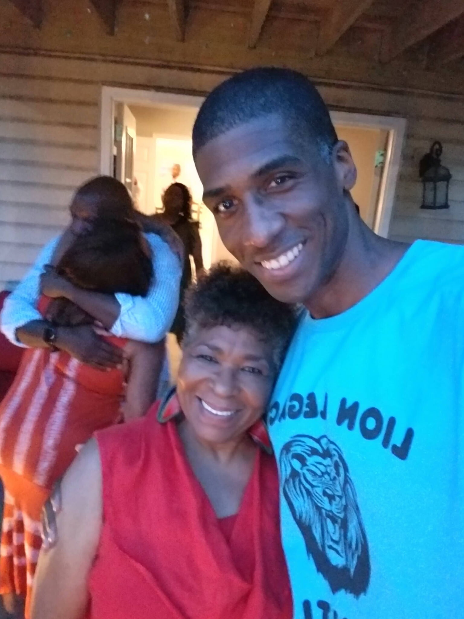 Smiling man in a blue shirt taking a selfie with an older woman in red on a porch, while others hug in the background.