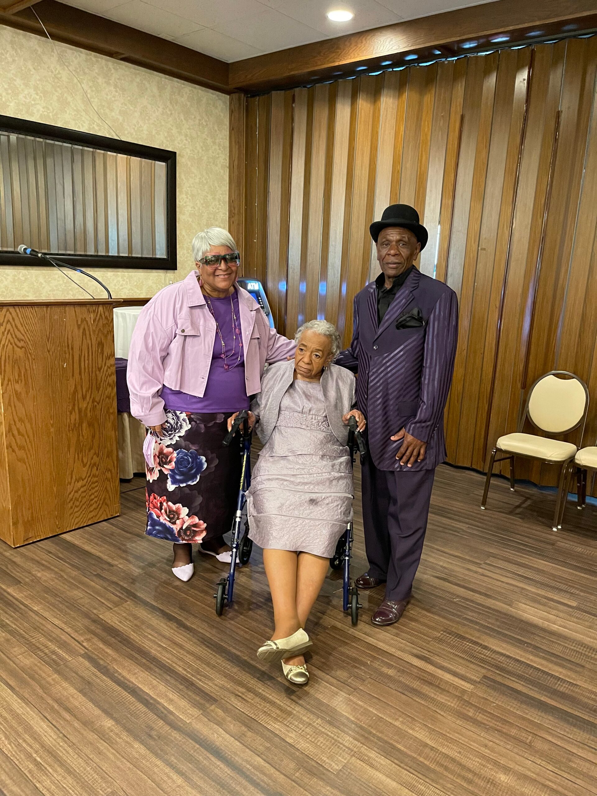 Three seniors posing together in an event hall: elderly woman in a wheelchair flanked by a smiling woman in lavender jacket and a man in a purple suit and hat, with a podium on the left.