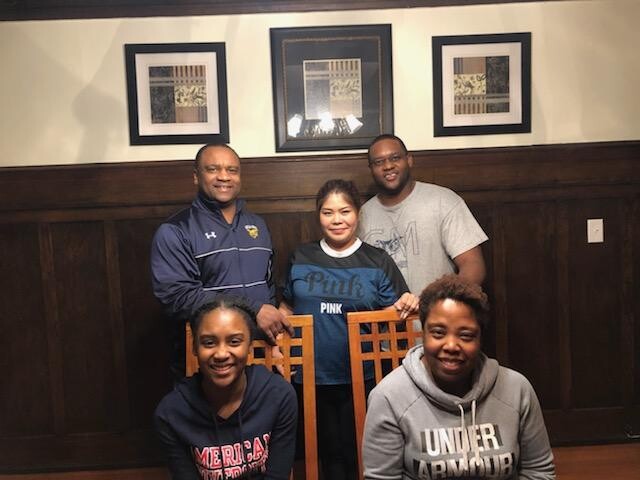 Five people posing for a casual photo in a dining area with dark wood paneling and framed artwork on the wall; two standing at the back and three seated in front, smiling at the camera.