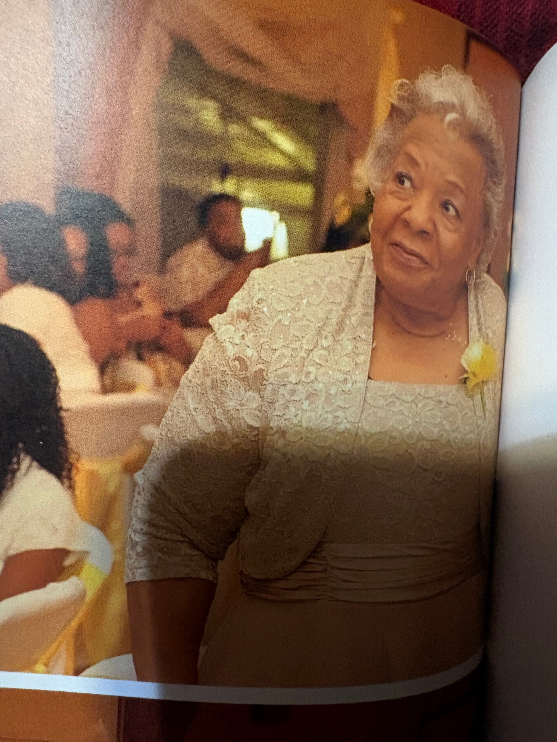 Older woman with short gray hair in a cream lace outfit, wearing a yellow corsage, looking to the side at a social event.