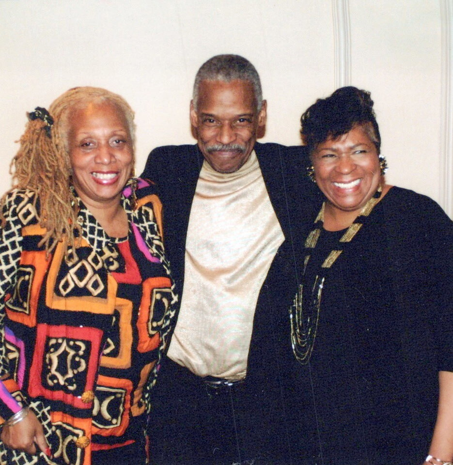 Three smiling adults posing together indoors, man in center wearing a beige turtleneck and blazer with two women on each side in colorful and black outfits.