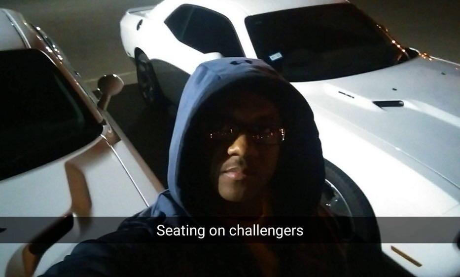 Person in a dark hoodie and glasses taking a nighttime selfie in a parking lot with white cars in the background.