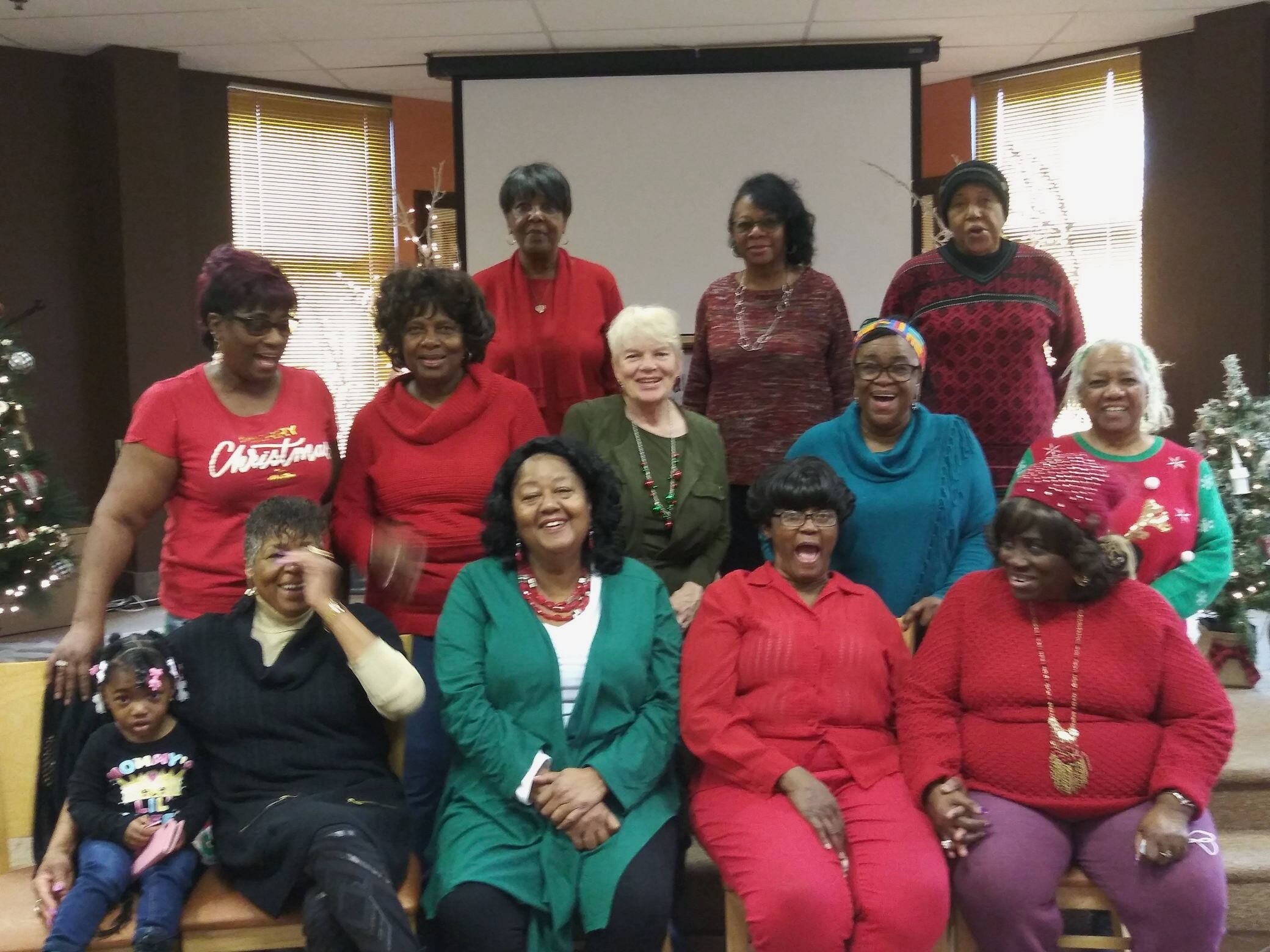 A multiethnic group of adults and children poses for a Christmas photo indoors, wearing festive red, green, and patterned sweaters.