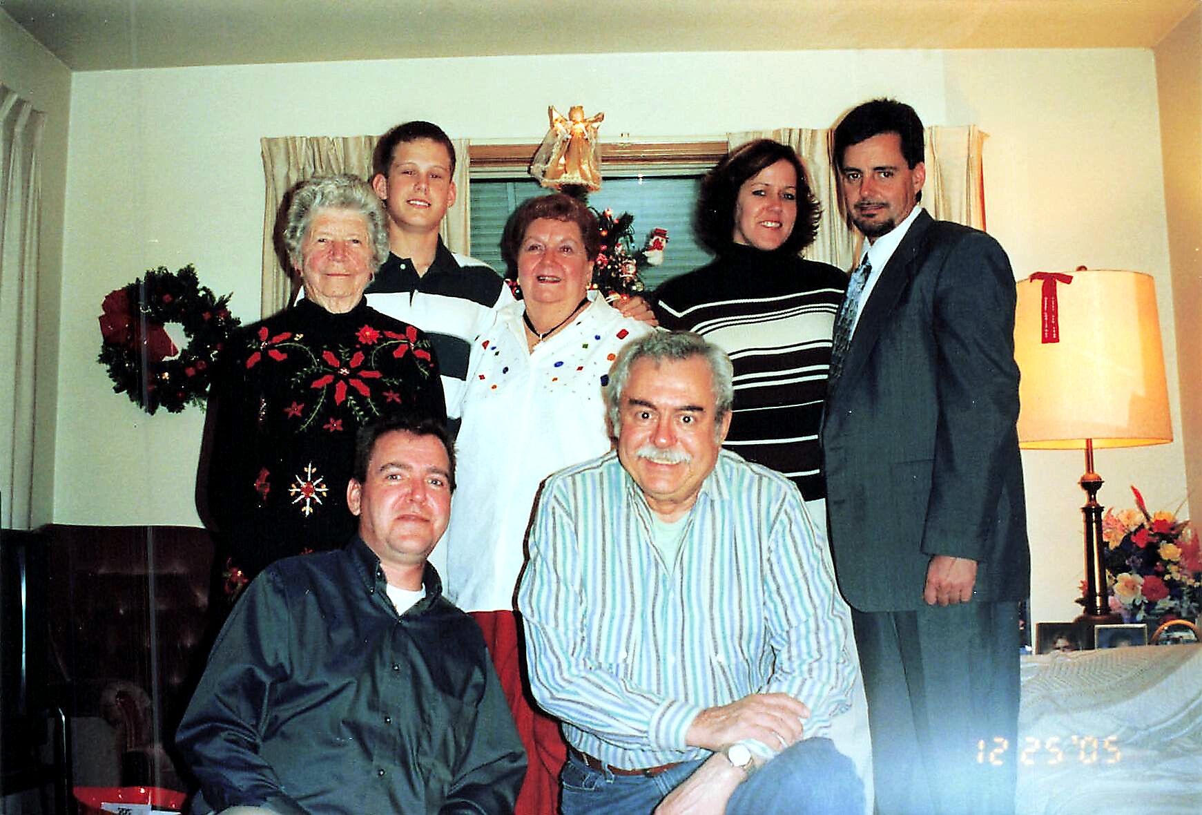 Group of seven people posing together in a living room for a Christmas photo, with a decorated tree and wreath in the background.