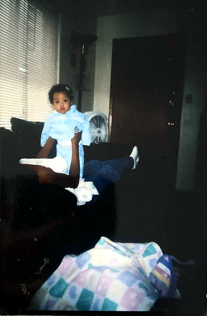 Baby in light-blue clothing held up by an adult in a living room; window blinds, a standing fan, and a closed door in the background.