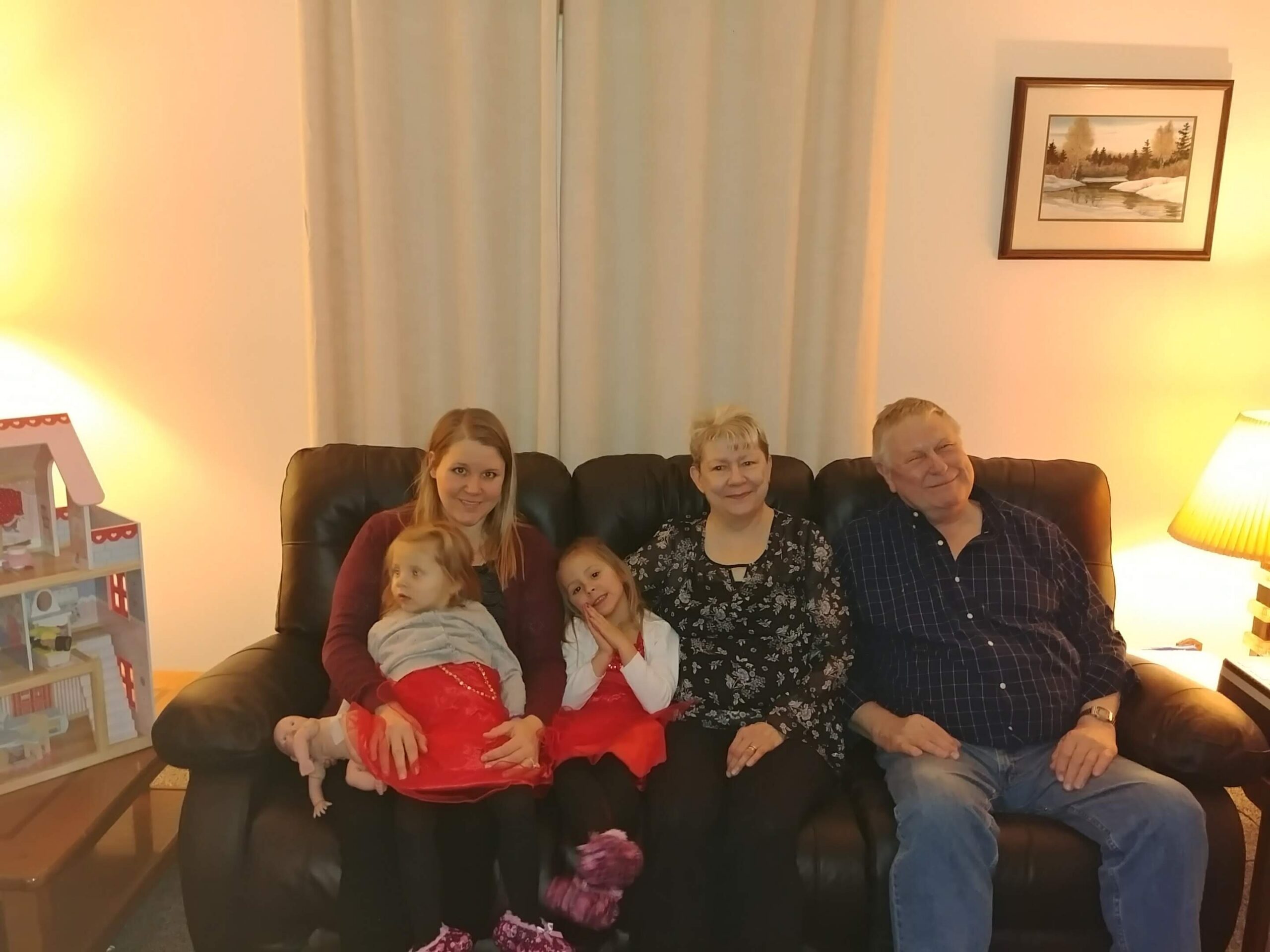 Family posing on a black leather sofa in a cozy living room; two young girls in red dresses with adults smiling nearby.